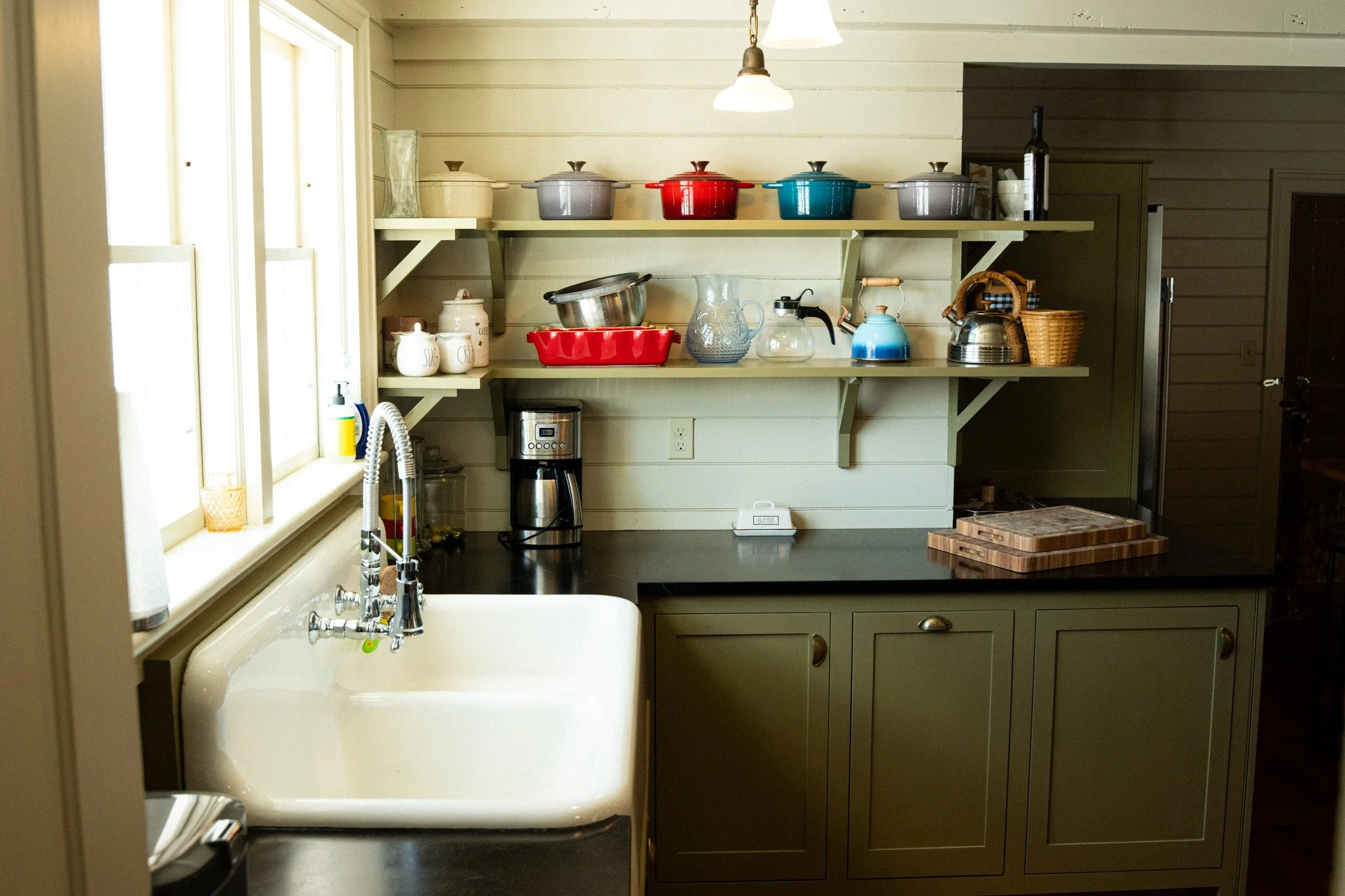 Kitchen with open shelves holding colorful pots and kitchenware, window by farmhouse sink, coffee maker, and cutting boards on dark countertop.