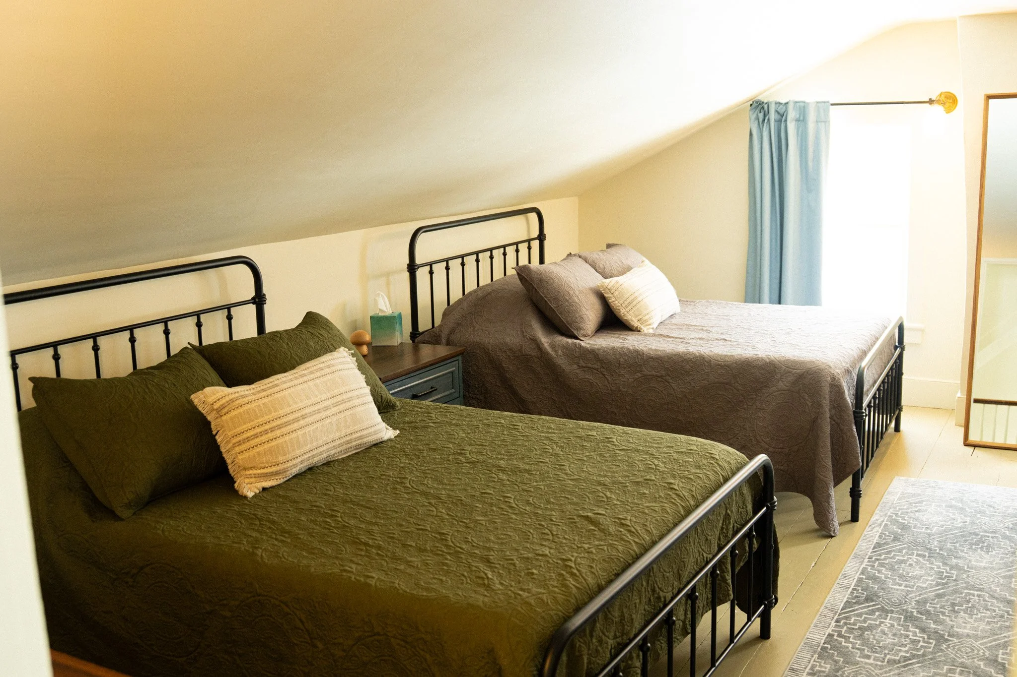 A cozy bedroom with two black metal beds, one with a green quilt and the other with a brown quilt, each with multiple pillows. A small blue nightstand with a tissue box and a lamp is between the beds. There is a window with light blue curtains and a 