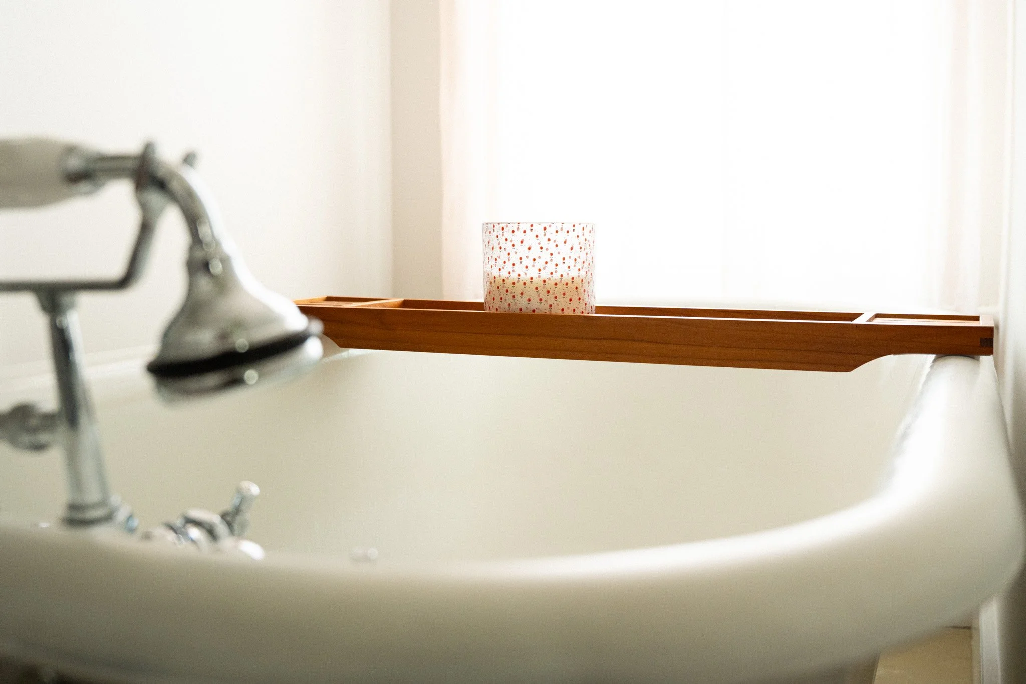 A white bathtub with silver fixtures, partially visible in the foreground, and a wooden tray holding a white candle with a dotted pattern, placed on the edge of the tub near a bright window.