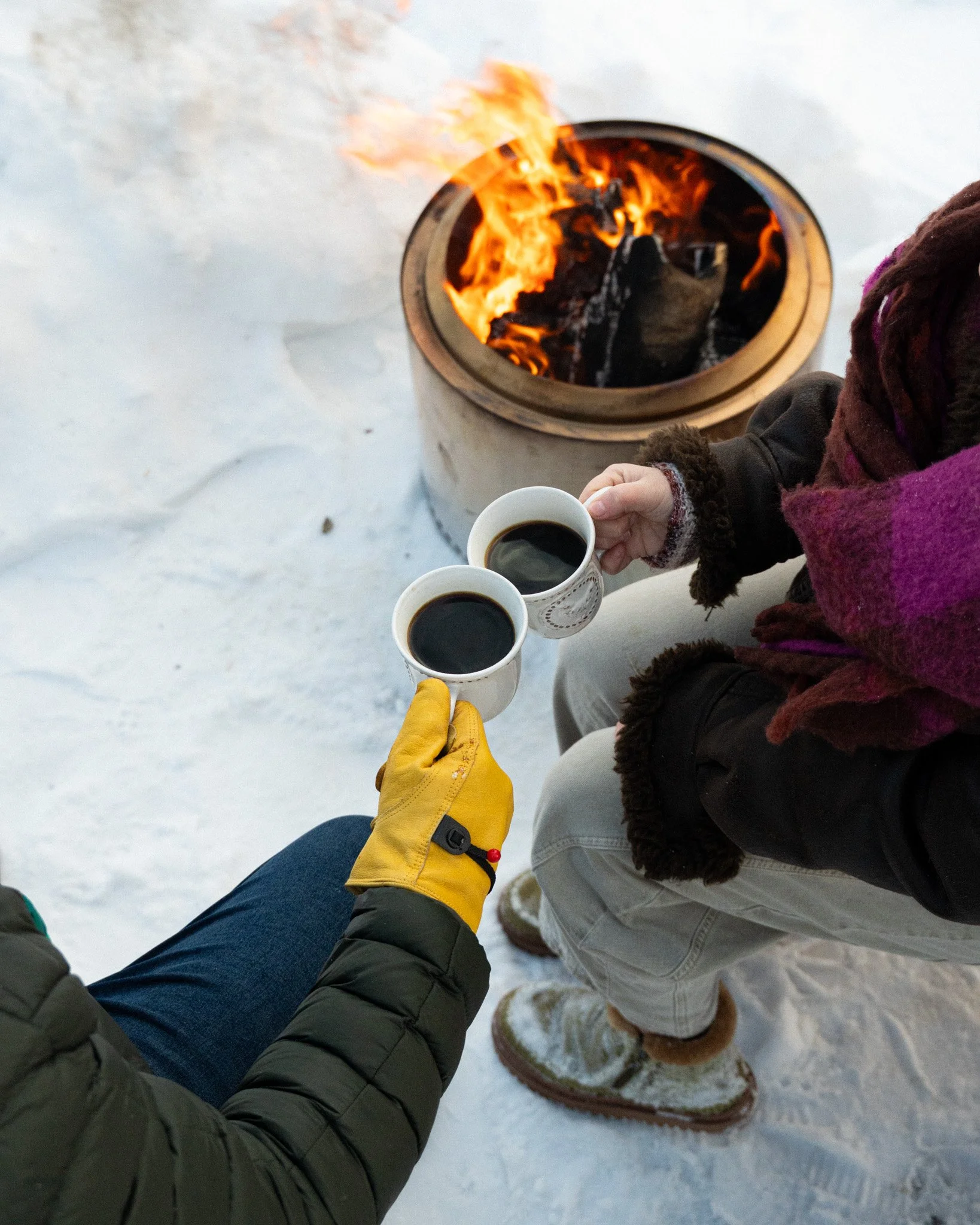 Two people holding cups of black coffee outdoors near a fire pit on snow, wearing winter clothing including gloves, scarves, and winter boots.