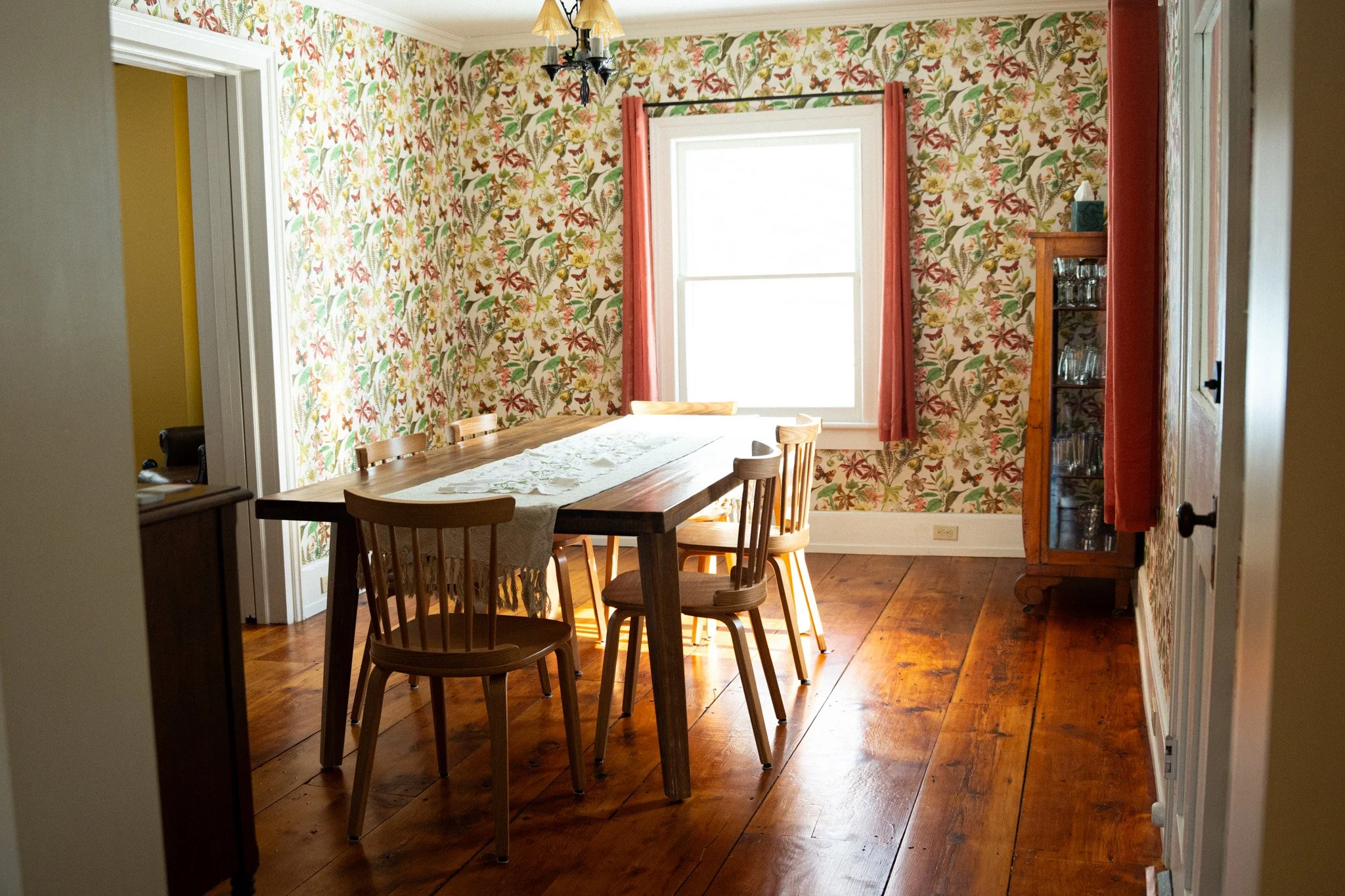 A dining room with floral wallpaper, a wooden table with a white lace table runner, and six wooden chairs. There is a window with coral curtains, and a wooden cabinet with glassware on the right side. The floor is hardwood.
