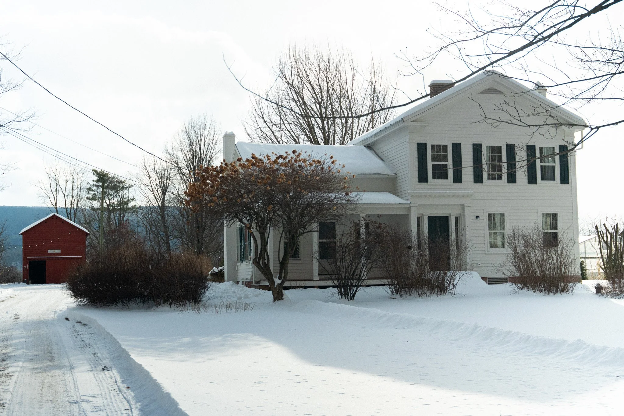 A white two-story house with black shutters in a snowy landscape with leafless trees and a red barn in the background.