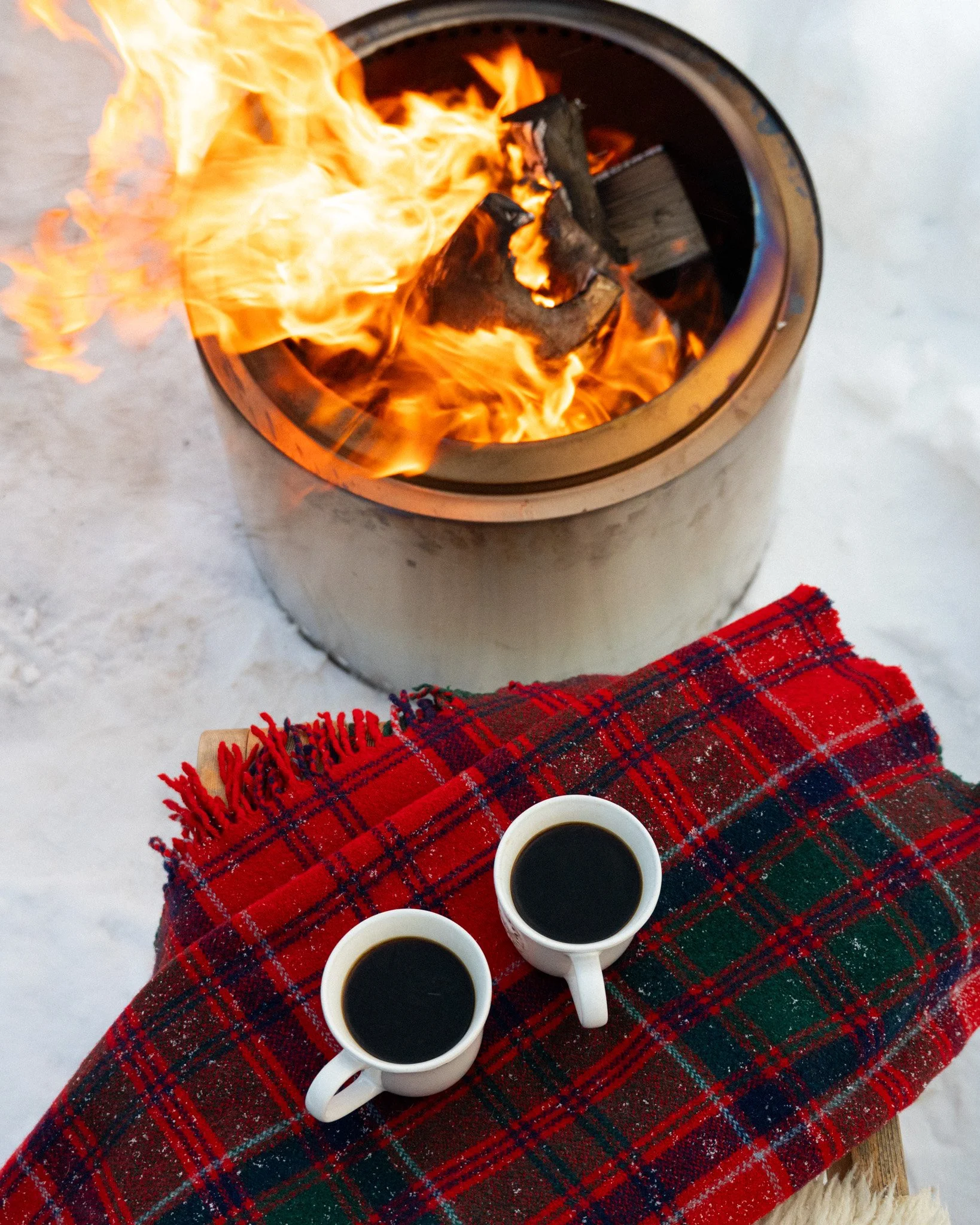Two cups of black coffee on a red tartan blanket next to a fire in a fire pit outdoors in a snowy setting.