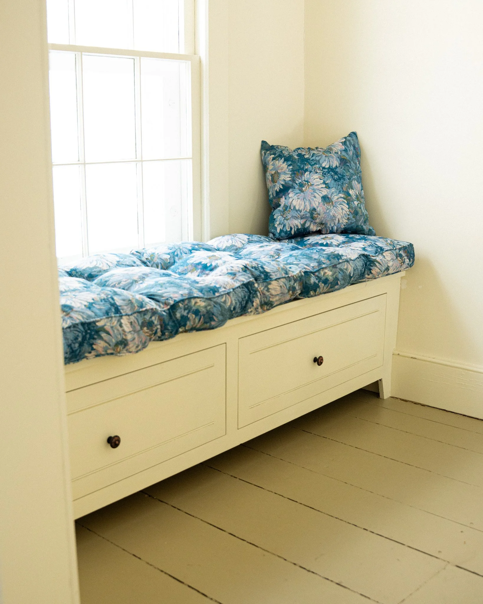 A window seat with floral blue and white cushions and a large matching pillow, built into a corner of a room with white walls and cream-colored wooden floors.