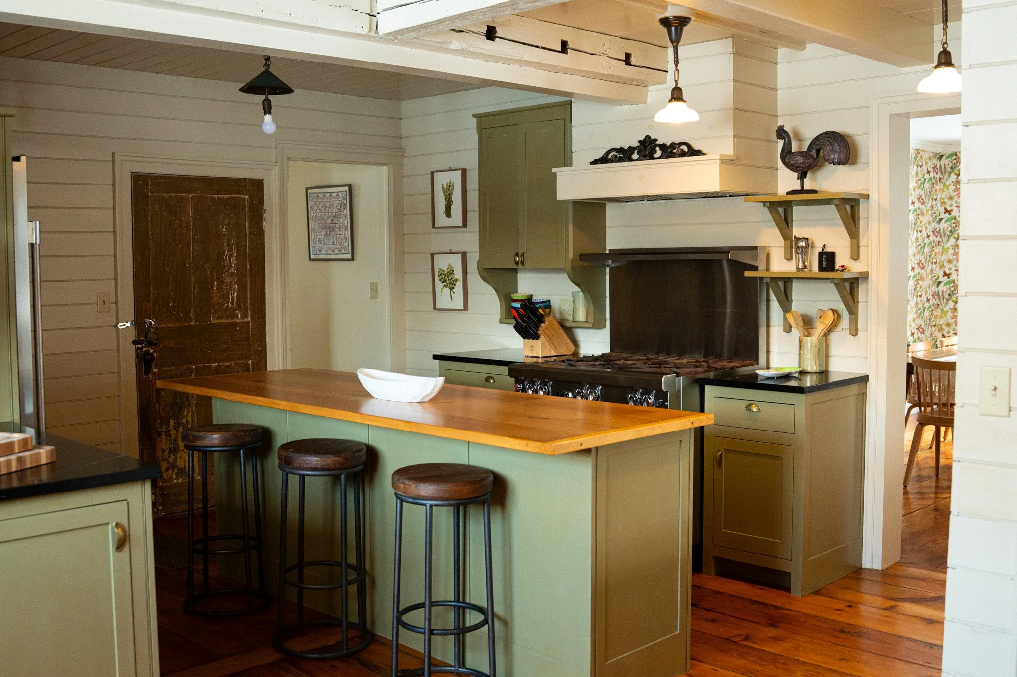Kitchen with green cabinets, wooden island with three stools, stove, and shelves with decorative items.