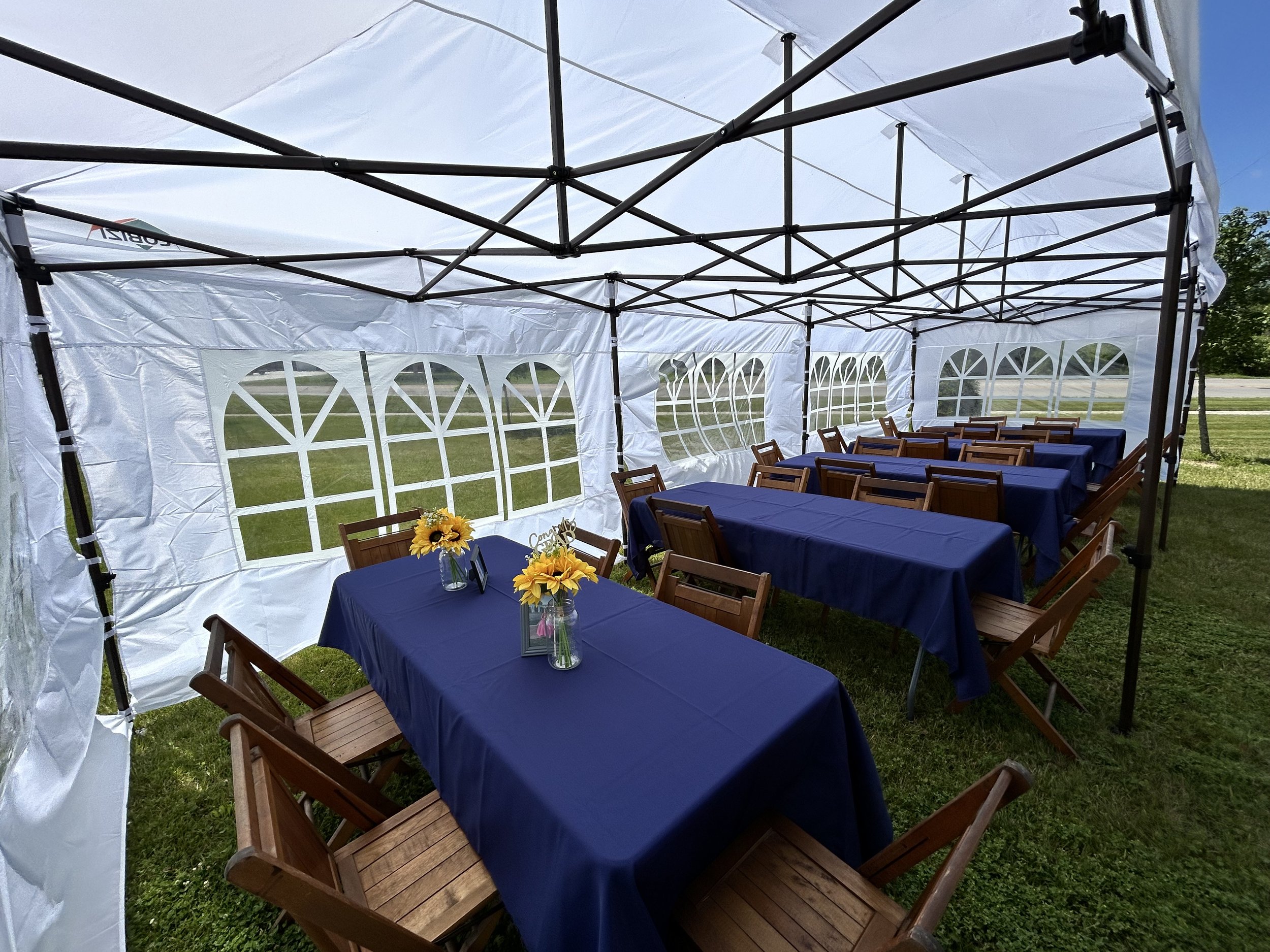 Outdoor event setup inside a white tent with blue tablecloths, wooden chairs, and sunflower centerpieces.