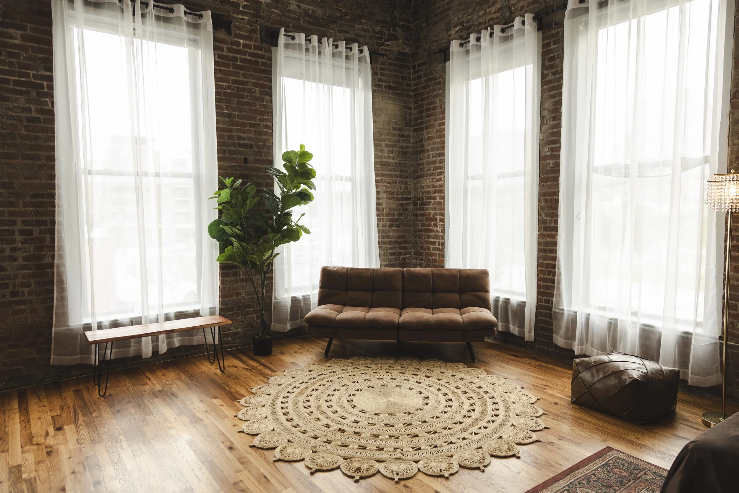 A green velvet chaise lounge in front of a large window in a room with exposed brick walls and wooden floors.