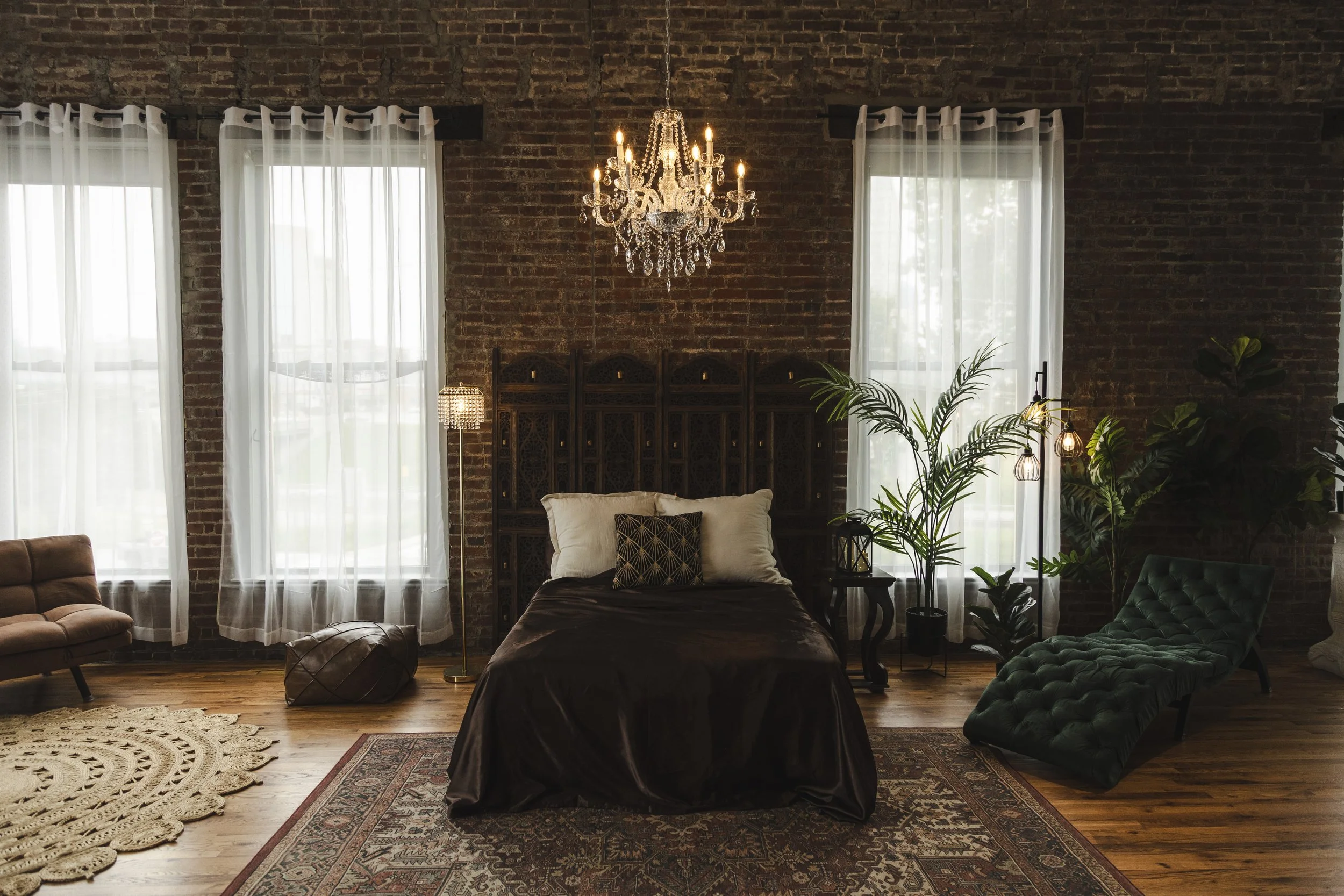 A modern, industrial-style living space with a dark wood floor, a gray velvet couch with pink pillows, a pink tufted ottoman, a hanging wicker chair with pillows, large windows letting in natural light, exposed brick walls, and a textured white wall.