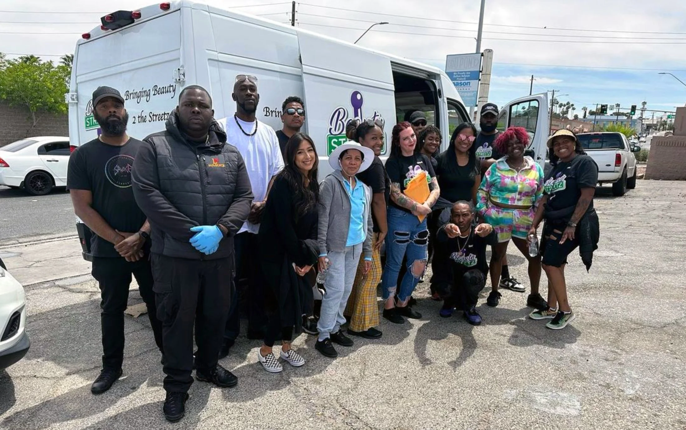 Group of diverse people gathered outdoors in front of a white van with a sign that says 'Bringing Beauty 2 the Streets'.