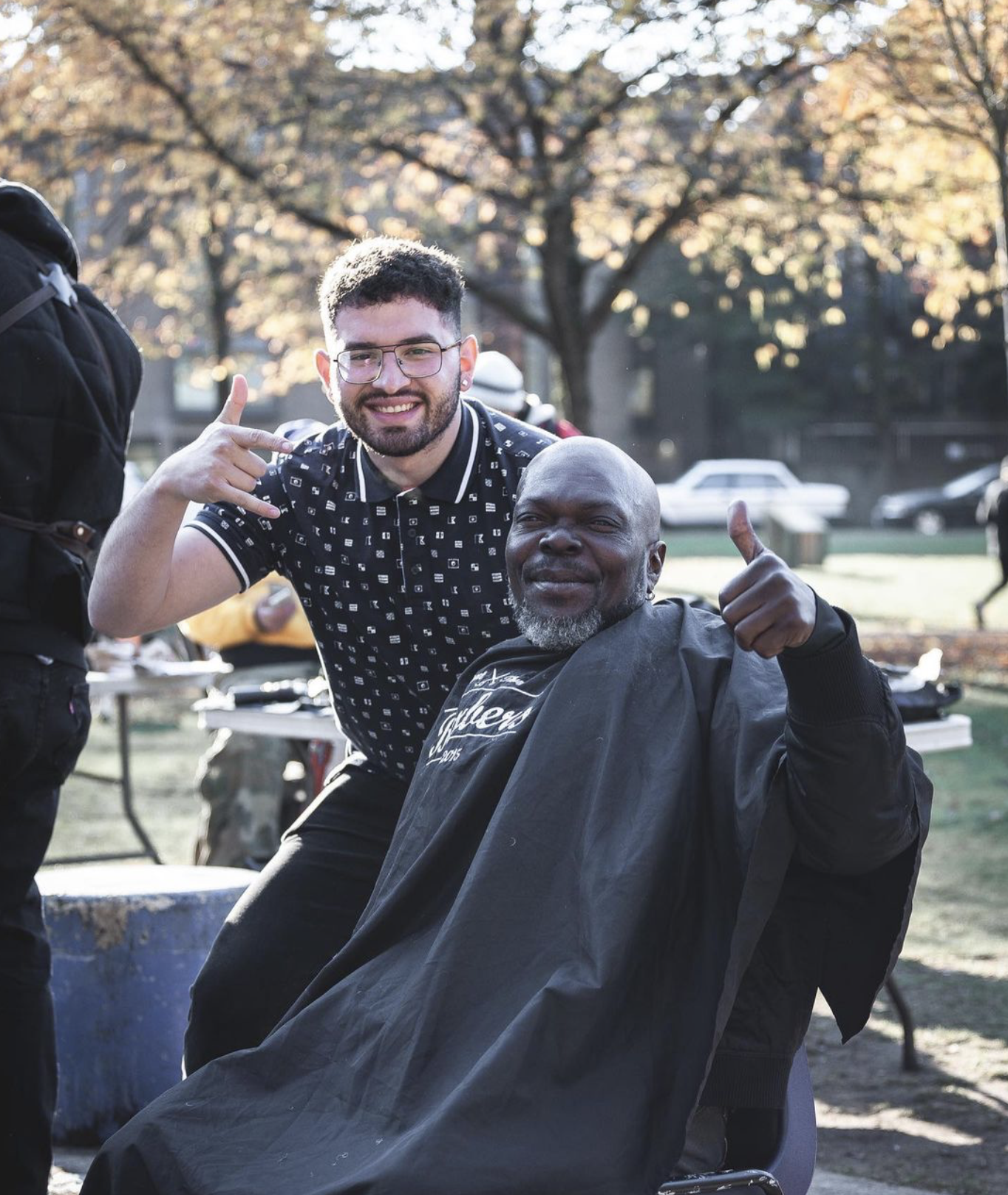 A young man with glasses giving a thumbs up and a middle-aged man with a beard smiling and making a hand gesture, outdoors with trees and cars in the background.