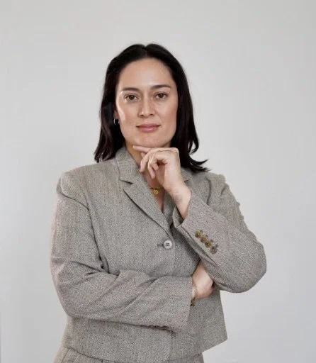Businesswoman with dark hair in a gray blazer, posing confidently with her hand near her chin against a plain gray background.
