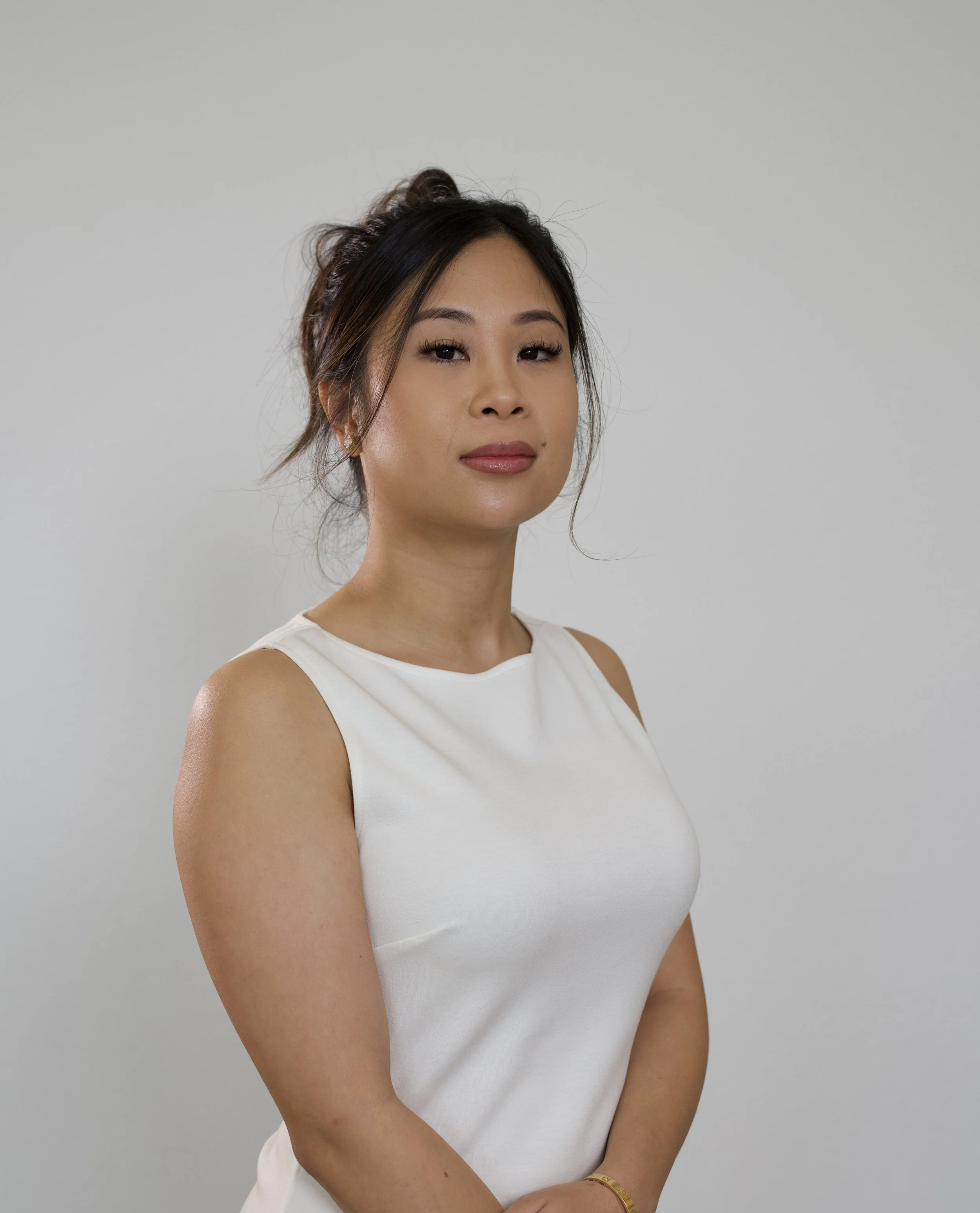 A woman with dark hair tied up, wearing a sleeveless white top, standing against a plain gray background.