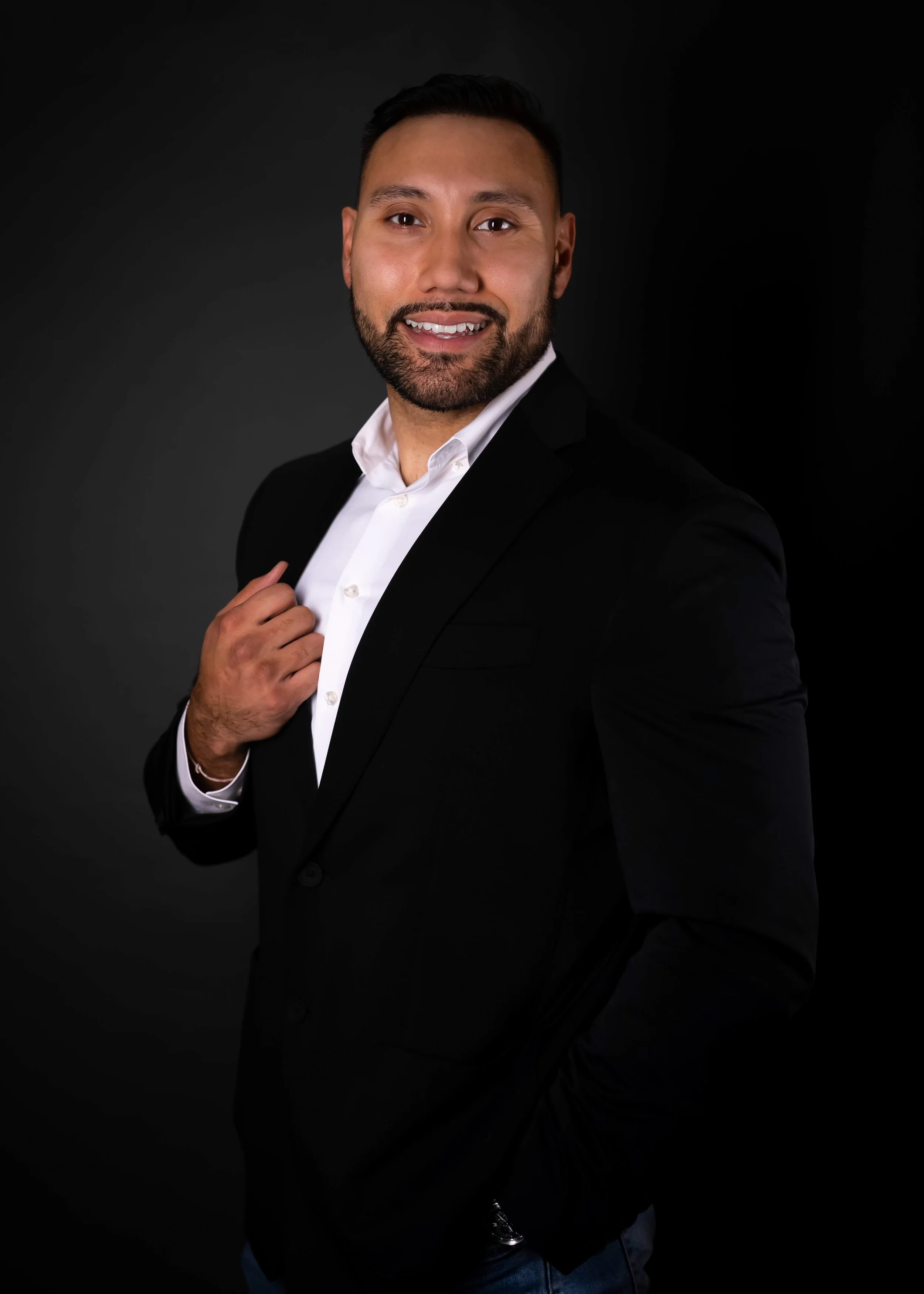 A man in a black suit jacket and white shirt smiling against a dark background, with one hand adjusting his jacket lapel.