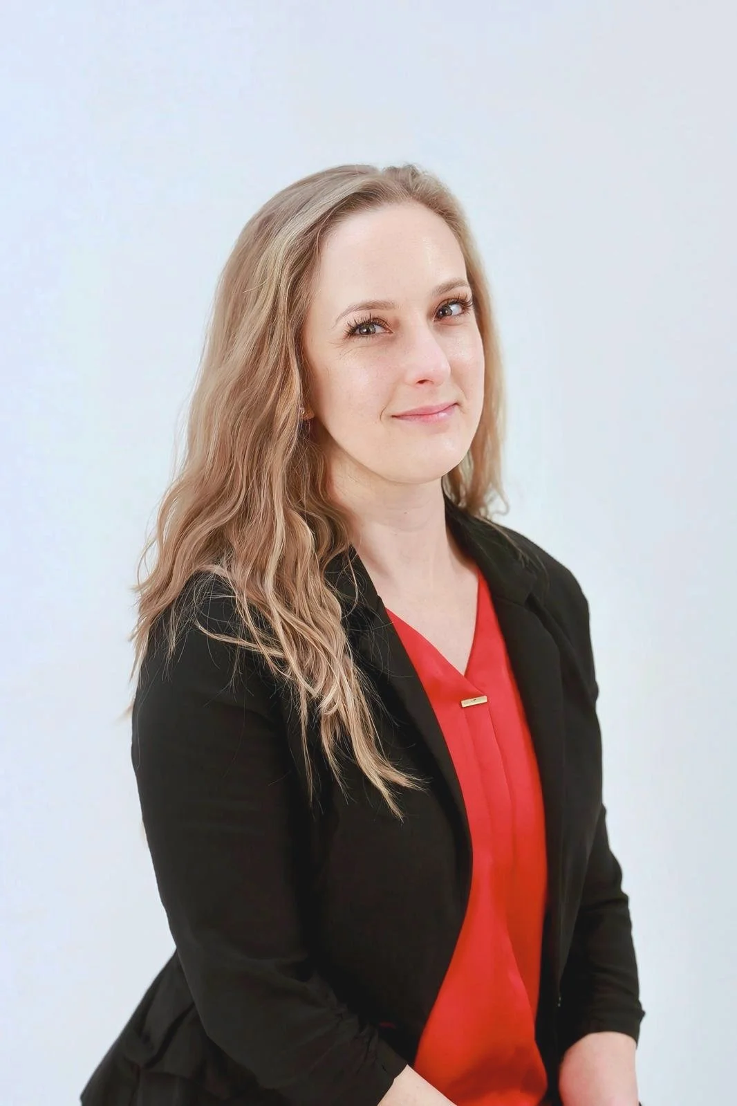 A woman with long wavy blonde hair, wearing a black blazer and a red top, standing against a plain light-colored background, smiling softly.