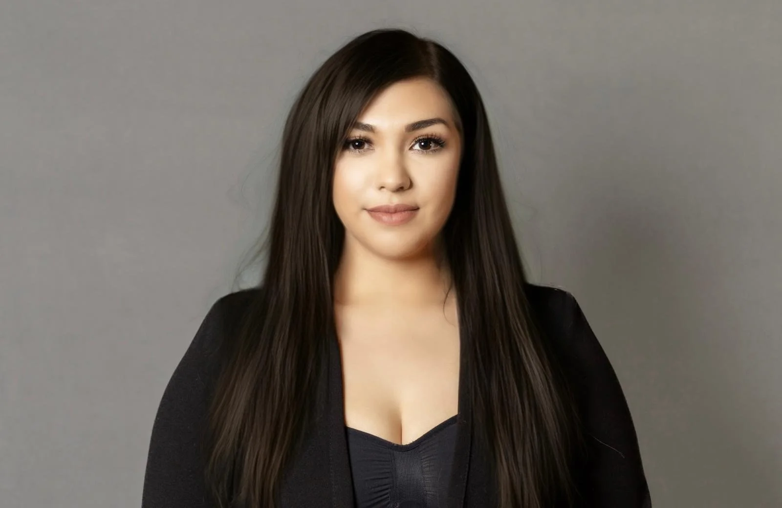 Portrait of a young woman with long dark hair wearing a black top and blazer, standing against a plain gray background.