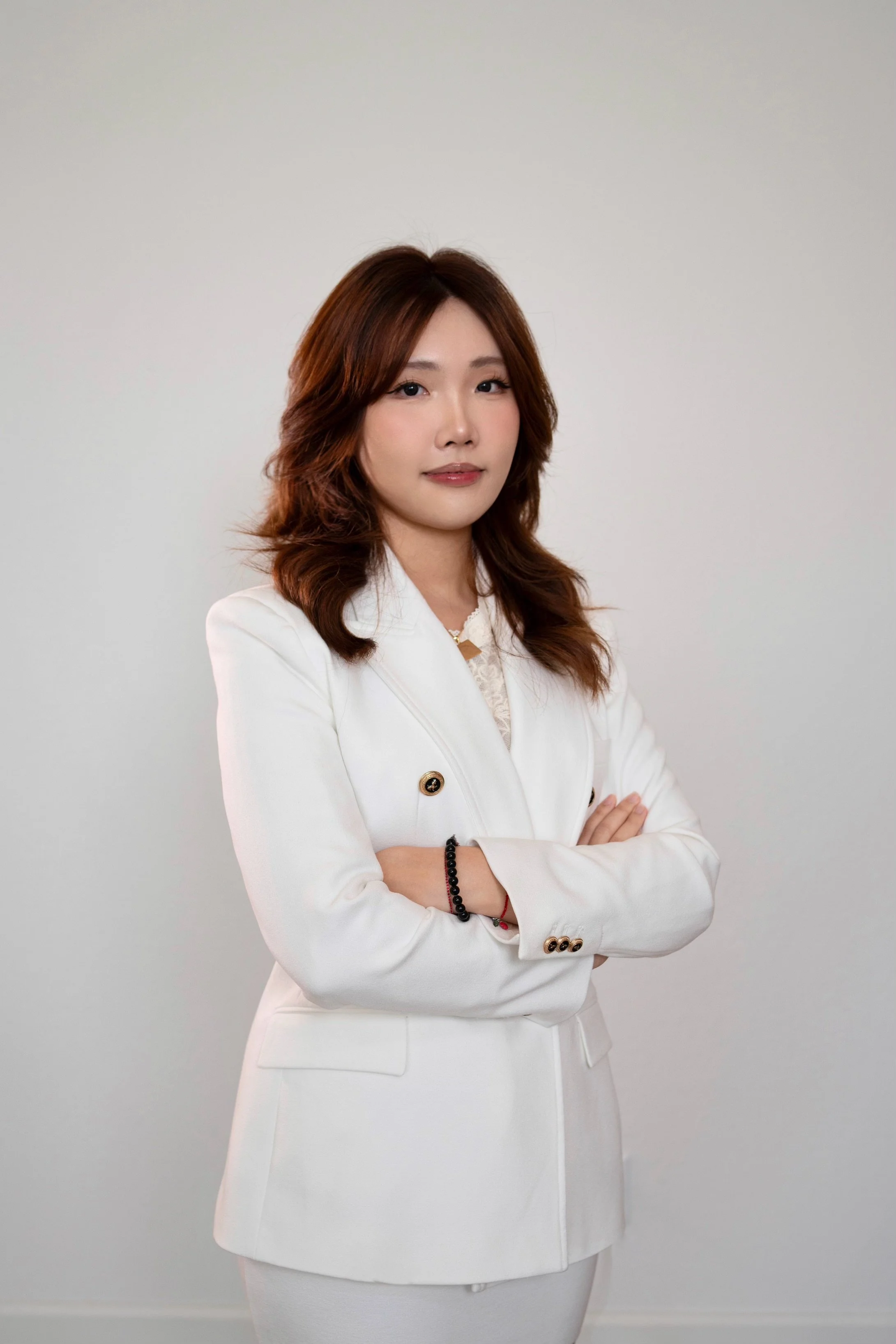 A woman with shoulder-length brown hair wearing a white blazer with arms crossed, standing against a plain white background.