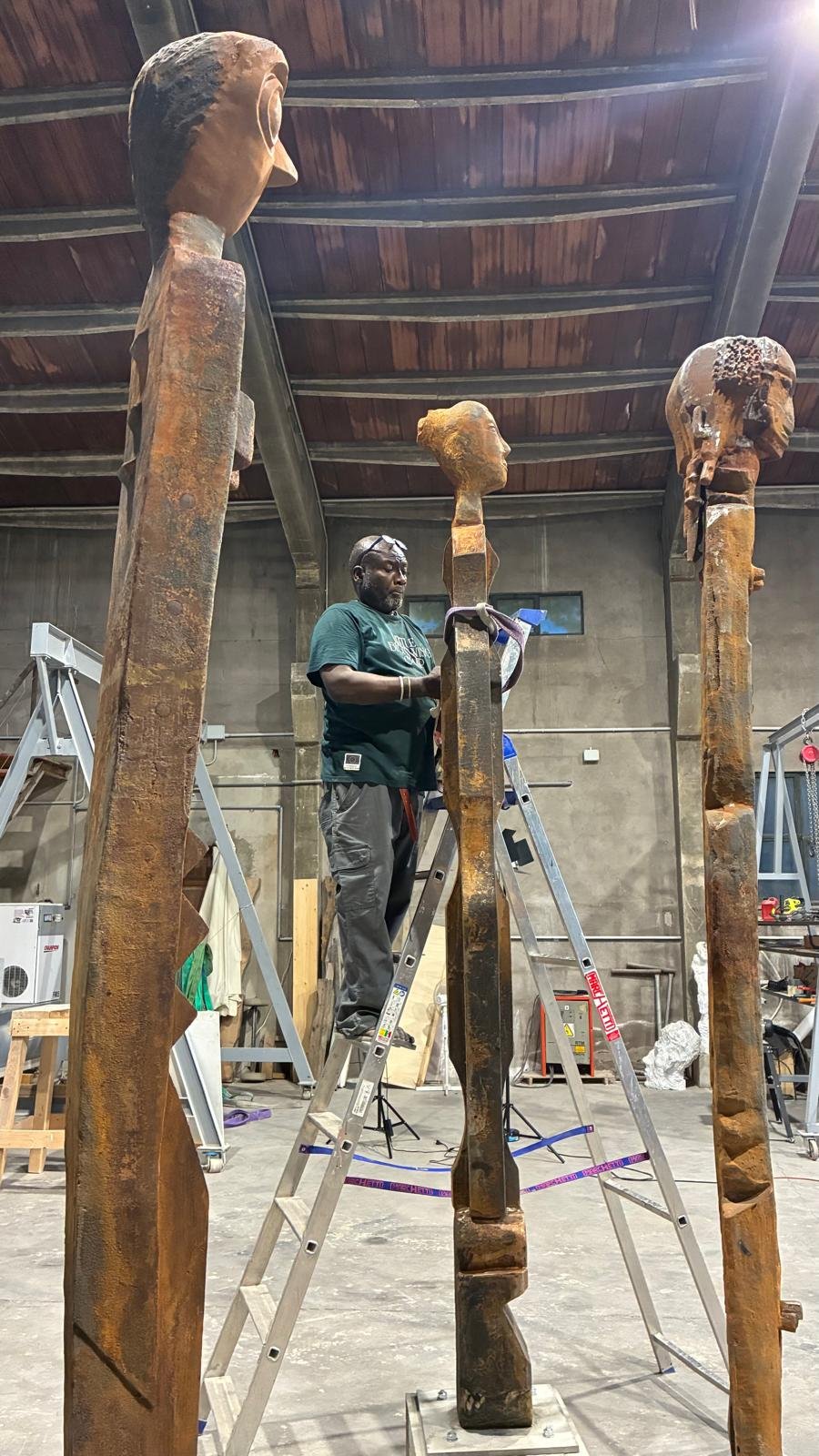 An artist working on a tall, wooden sculpture of a human head, using a ladder indoors with a high ceiling and exposed beams.