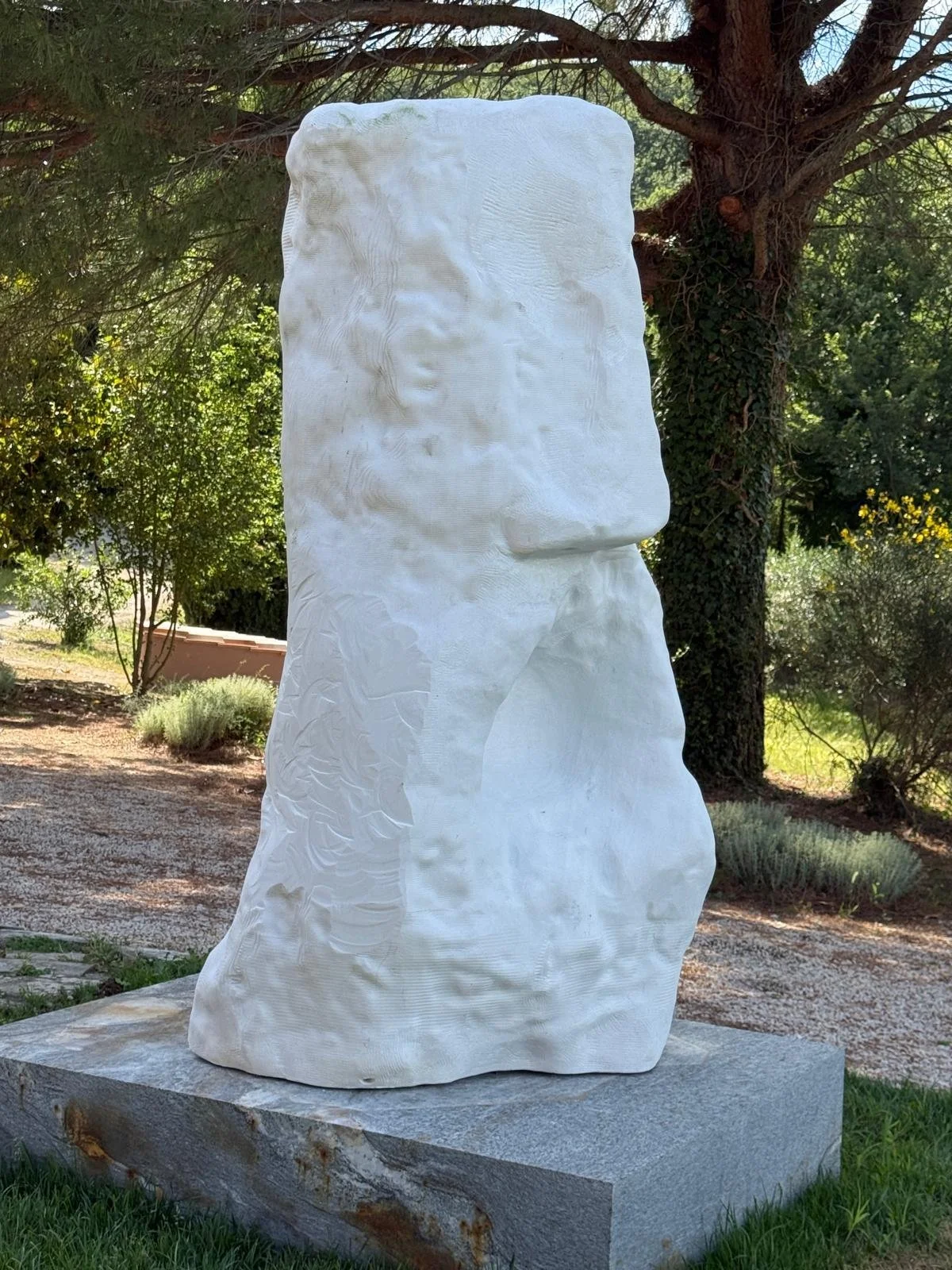 A white stone sculpture of a human face with minimal features, outdoors in a garden setting with trees and bushes in the background.