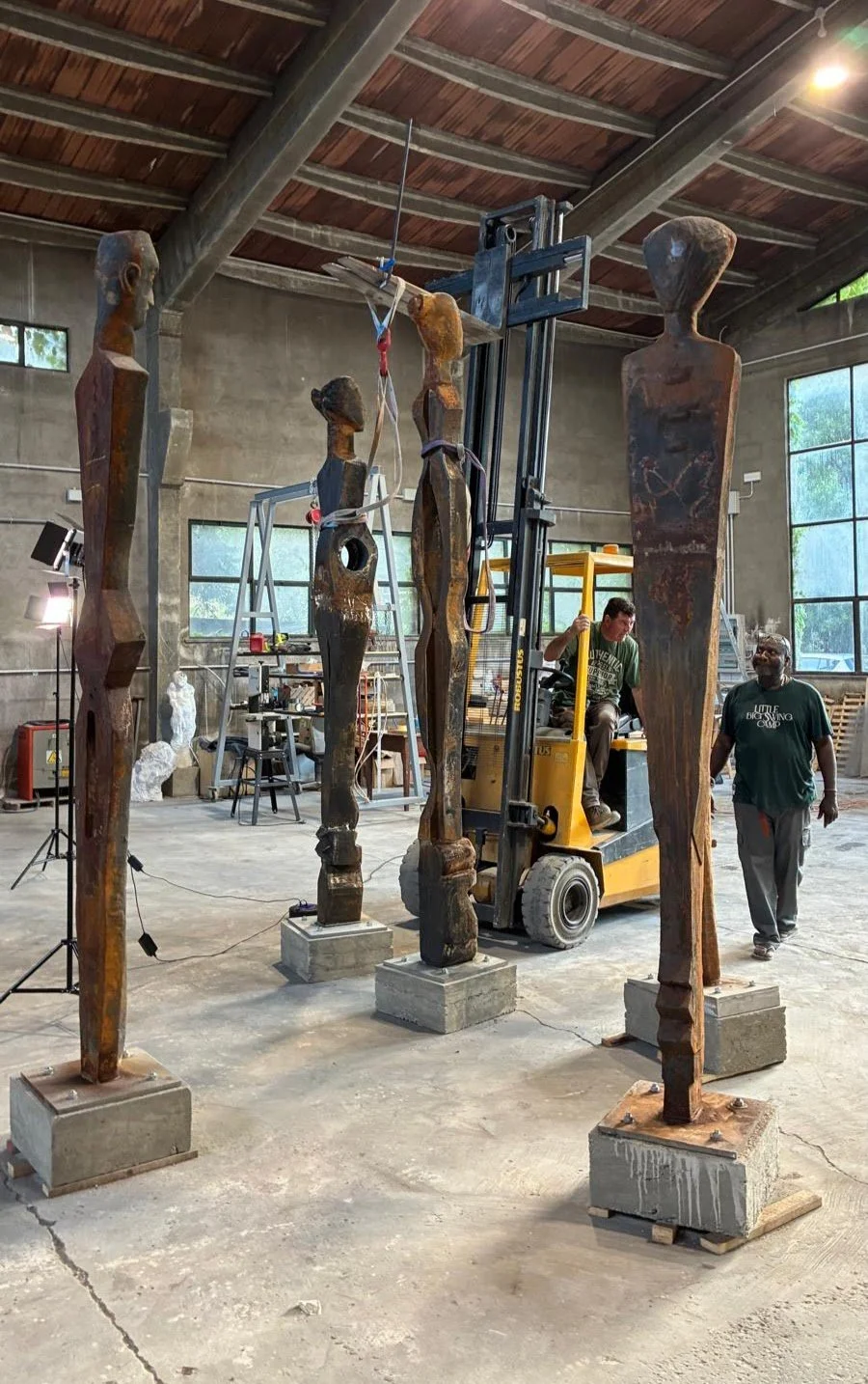 Four tall, abstract wooden sculptures in a workshop, with one being lifted by a forklift, two people working around them, and workshop tools in background.