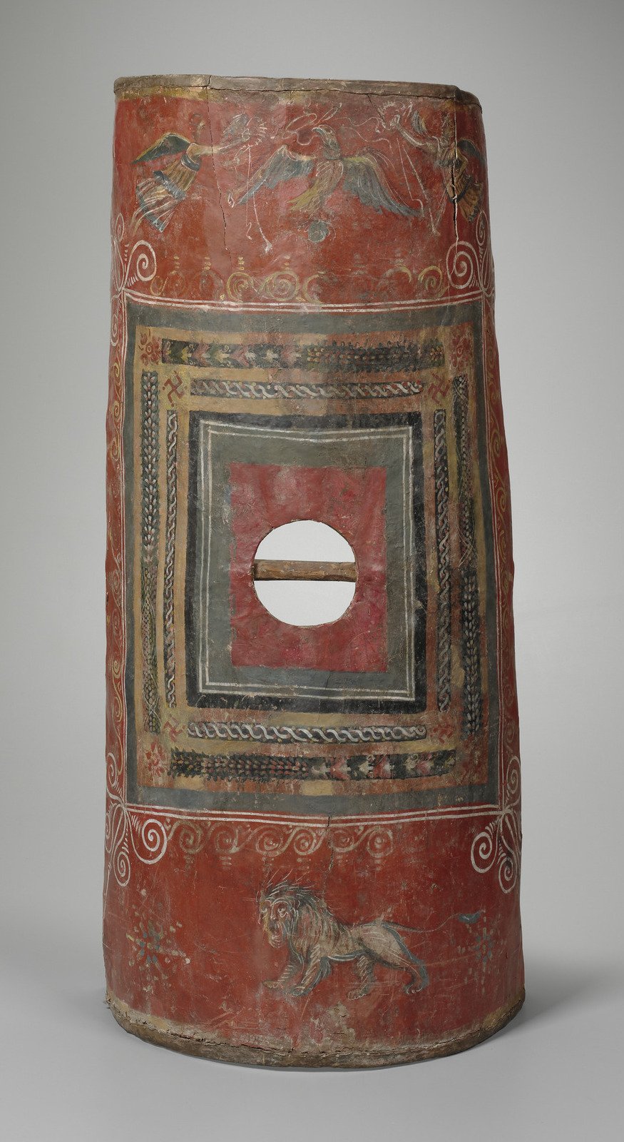 Ancient decorated shield with geometric patterns, animal figures including a lion, and mythological creatures on a red background.