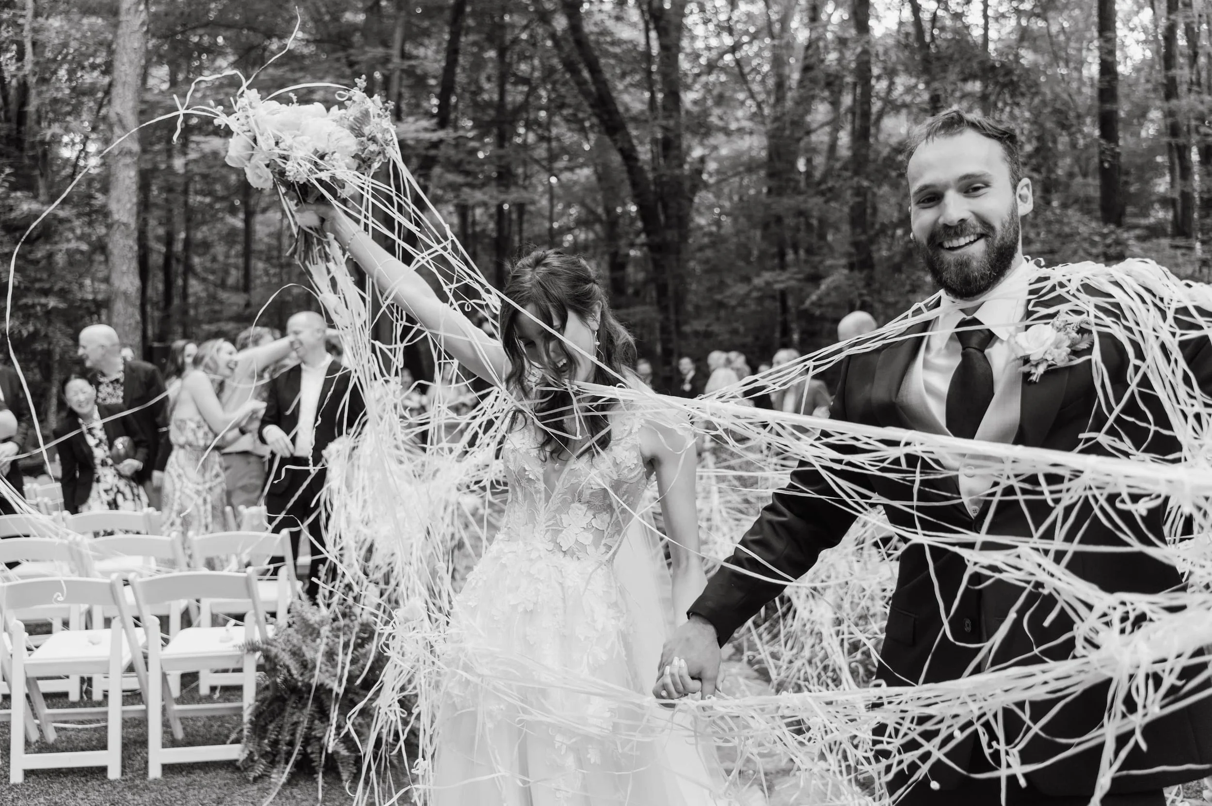 A wedding celebration outdoors in a wooded area. The bride and groom are holding hands through decorative ribbons, smiling, surrounded by guests. The image is in black and white.