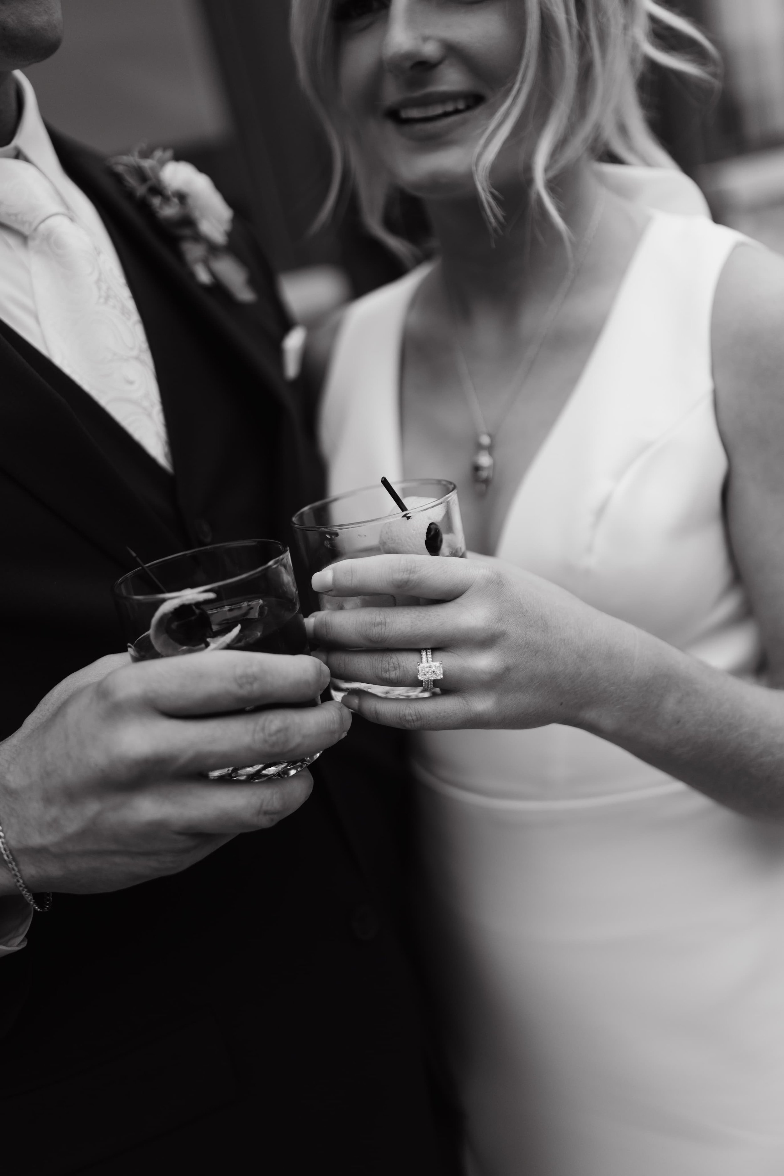 A close-up of a couple holding drinks during a celebration, with the woman's hand featuring an engagement ring.