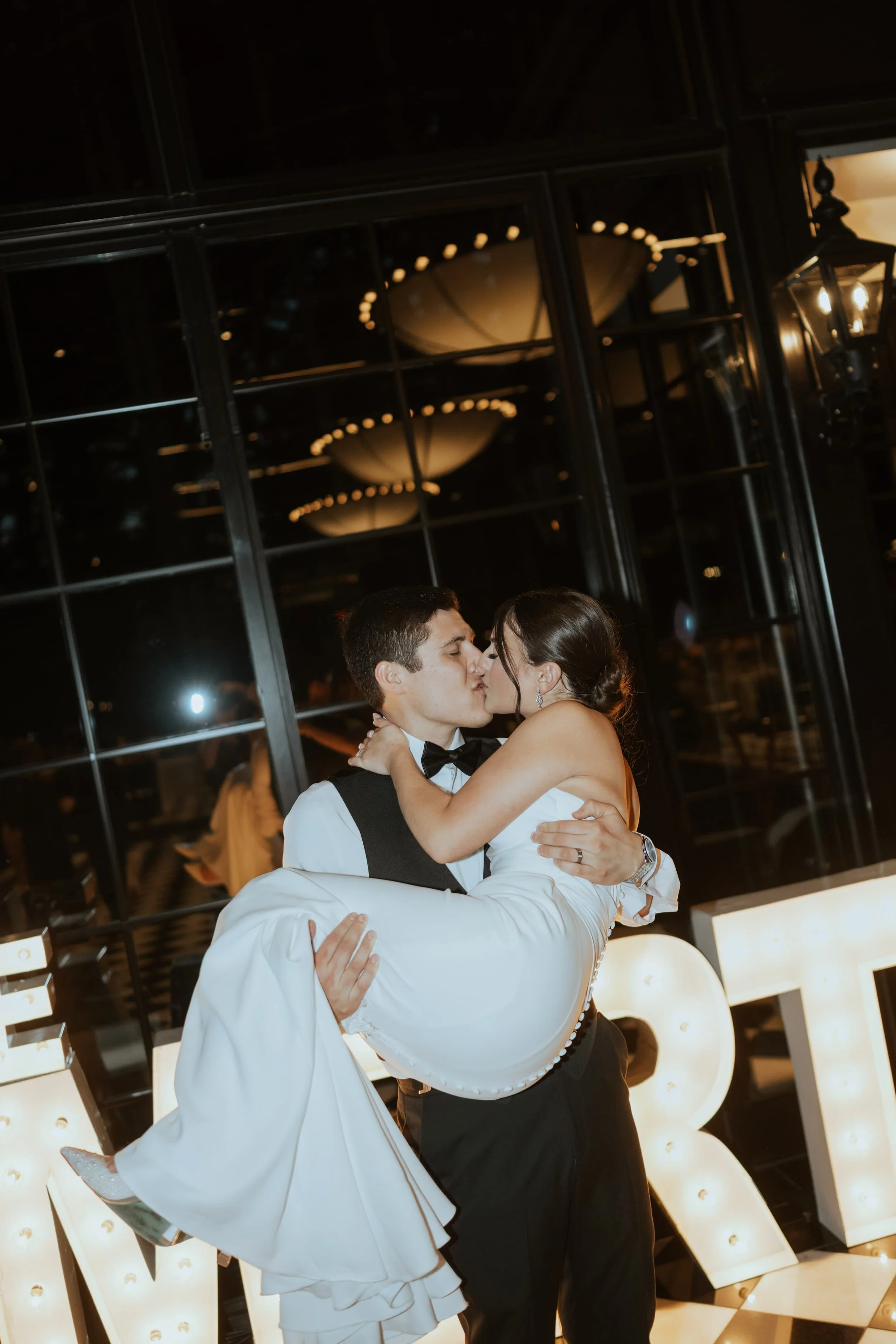 A man in tuxedo carrying a woman in a wedding dress as they kiss, with large illuminated letter signs in the background.