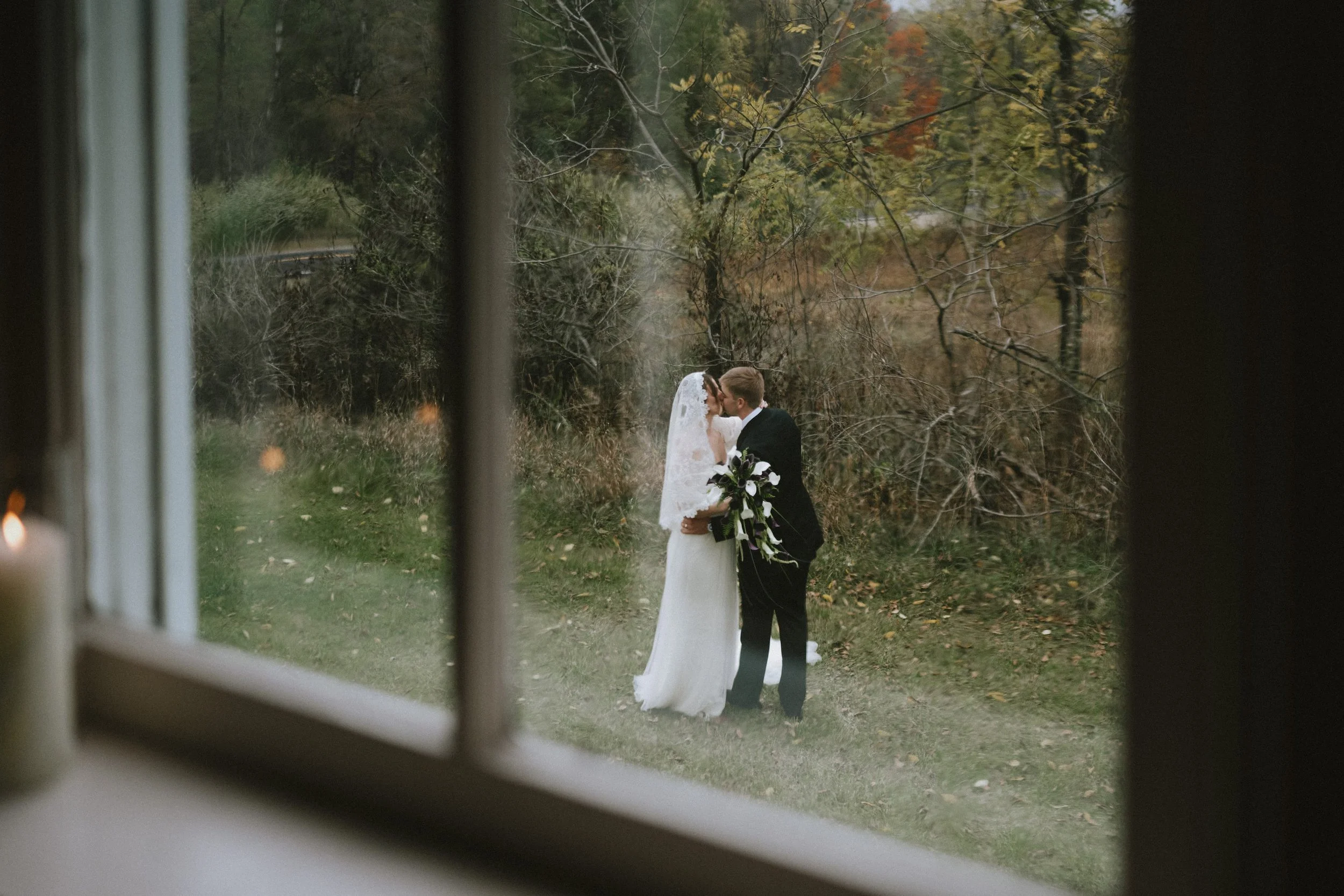 A bride and groom kissing outdoors in fall, viewed from inside through a window with candles on the sill.