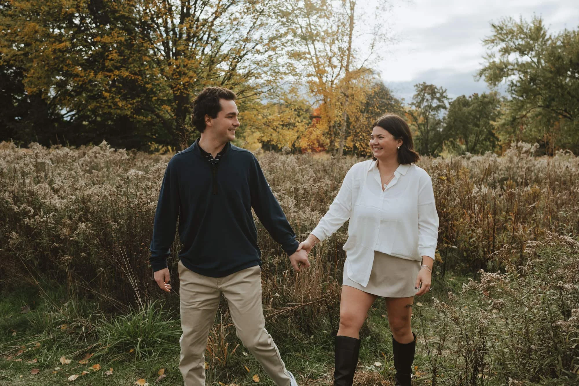 A young man and woman holding hands and walking outdoors in a field with fall foliage in the background, smiling at each other.