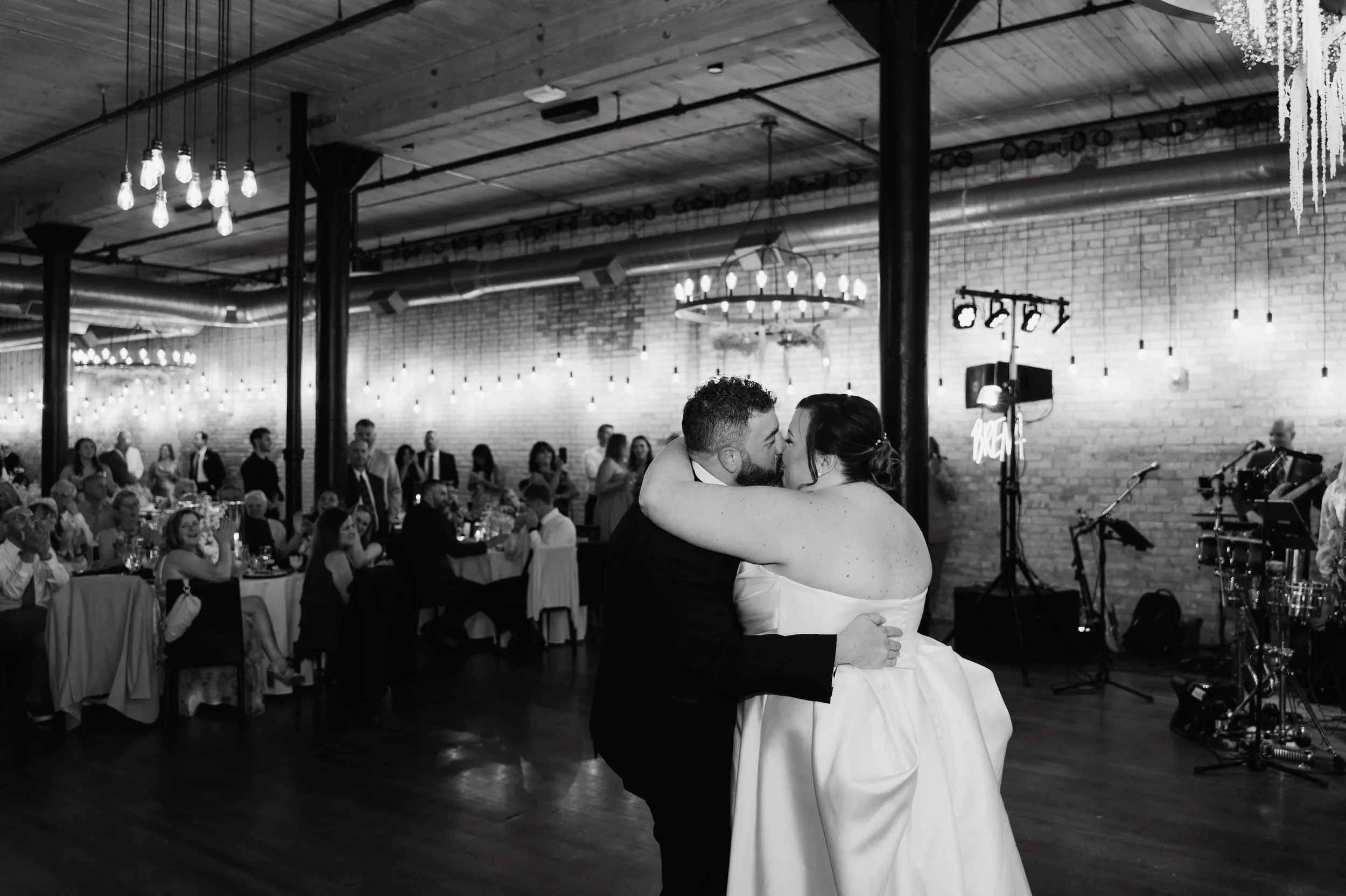 Black and white photo of a newlywed couple sharing a kiss while dancing at their wedding reception, with guests seated at tables and a band playing in the background.