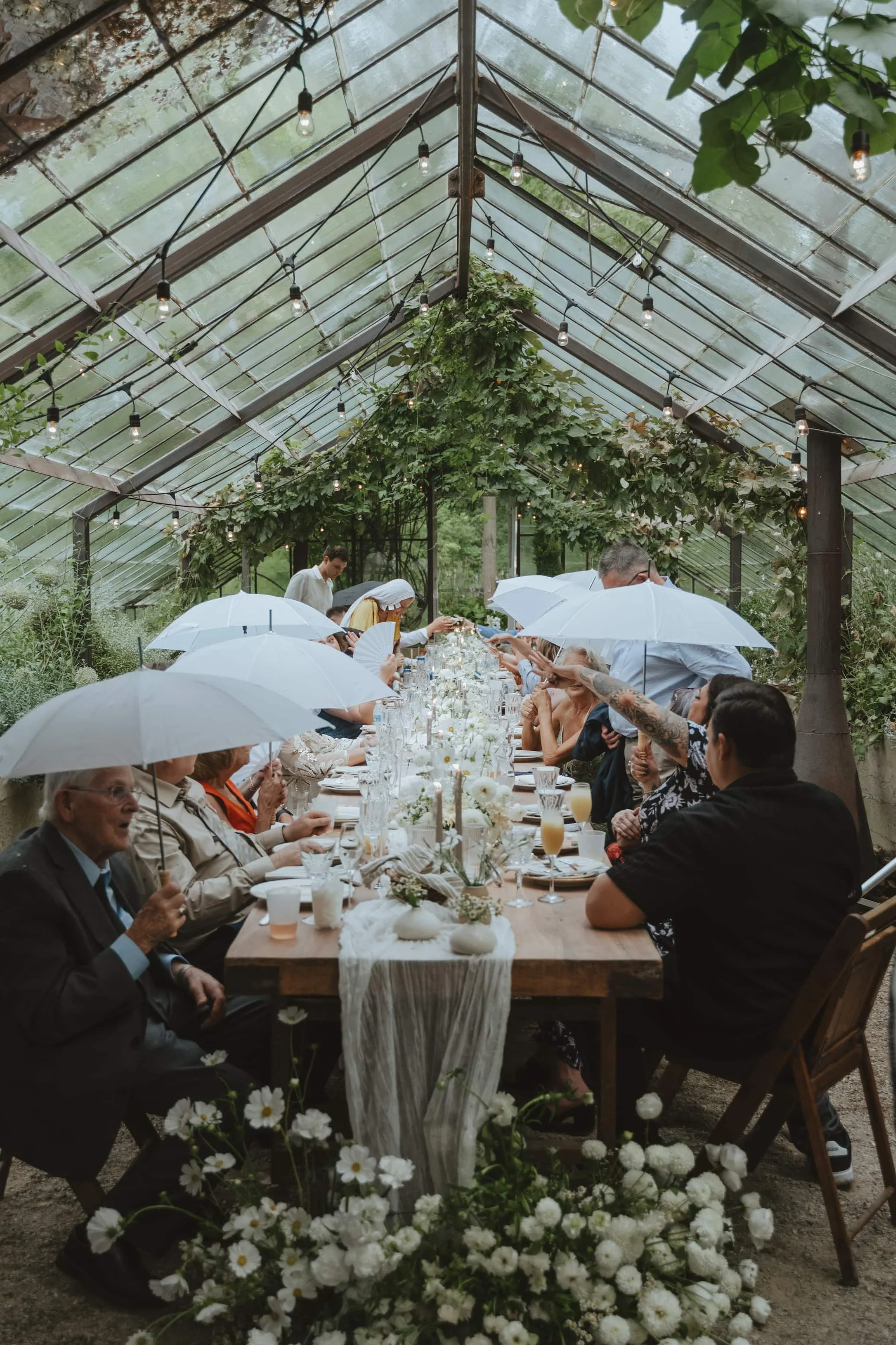 People gathered for a meal under a greenhouse canopy with string lights, holding white umbrellas, surrounded by greenery and floral decorations. Glass House Community located in Zeeland Michigan