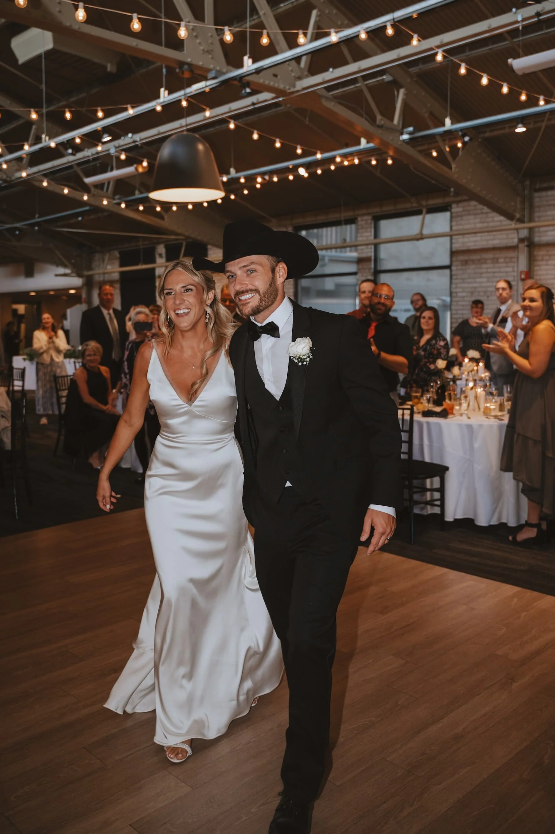 A bride and groom walking together at their wedding reception. The bride is wearing a satin white wedding gown, and the groom is in a black tuxedo with a cowboy hat. Guests are clapping and smiling in the background.