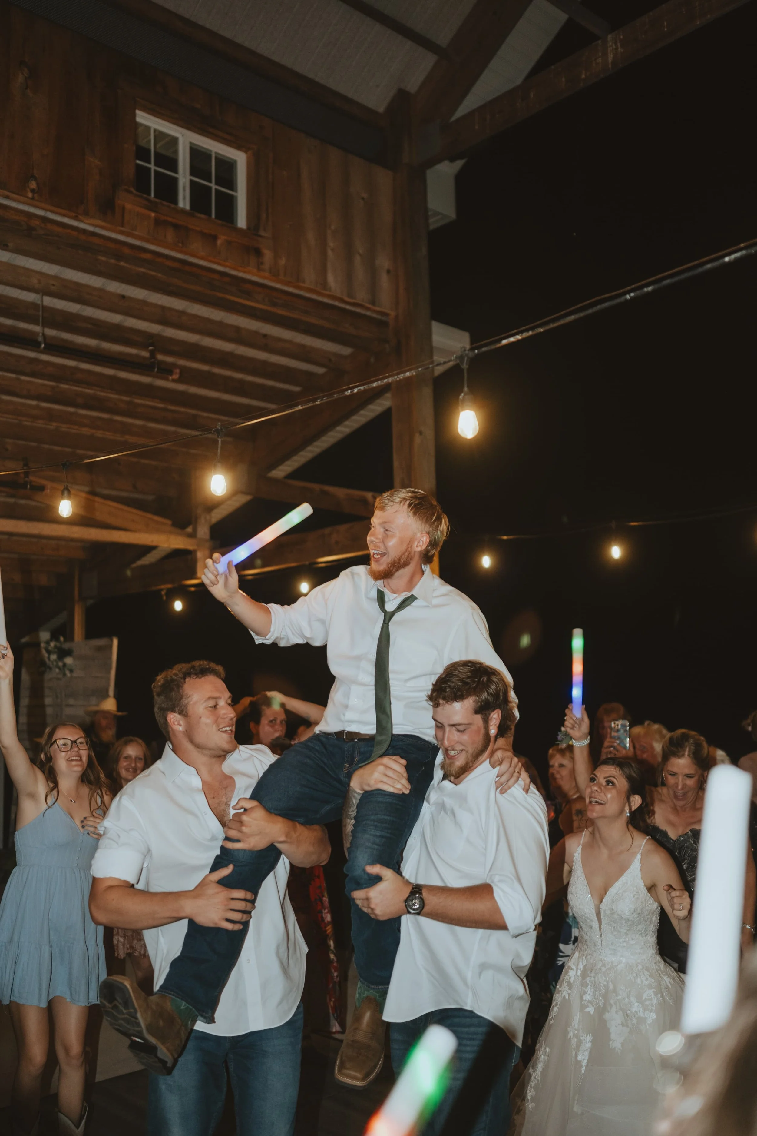 A group of people celebrating at a wedding reception, with a man being lifted into the air, holding a glow stick, and others smiling and dancing around him.