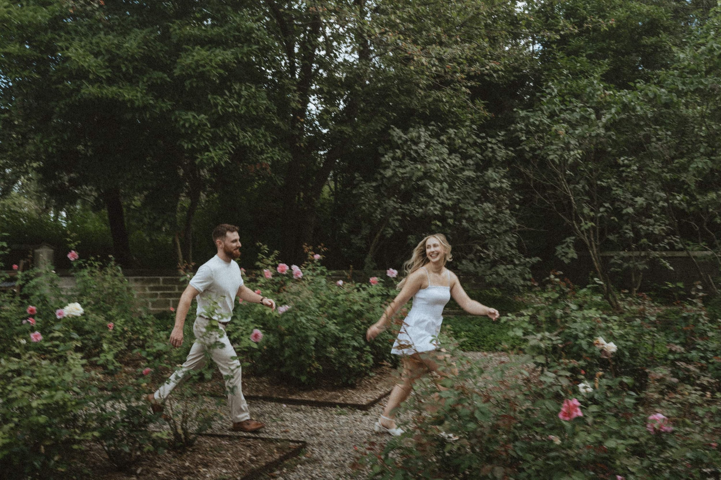 A joyful couple running through a garden with pink roses and tall trees in the background.