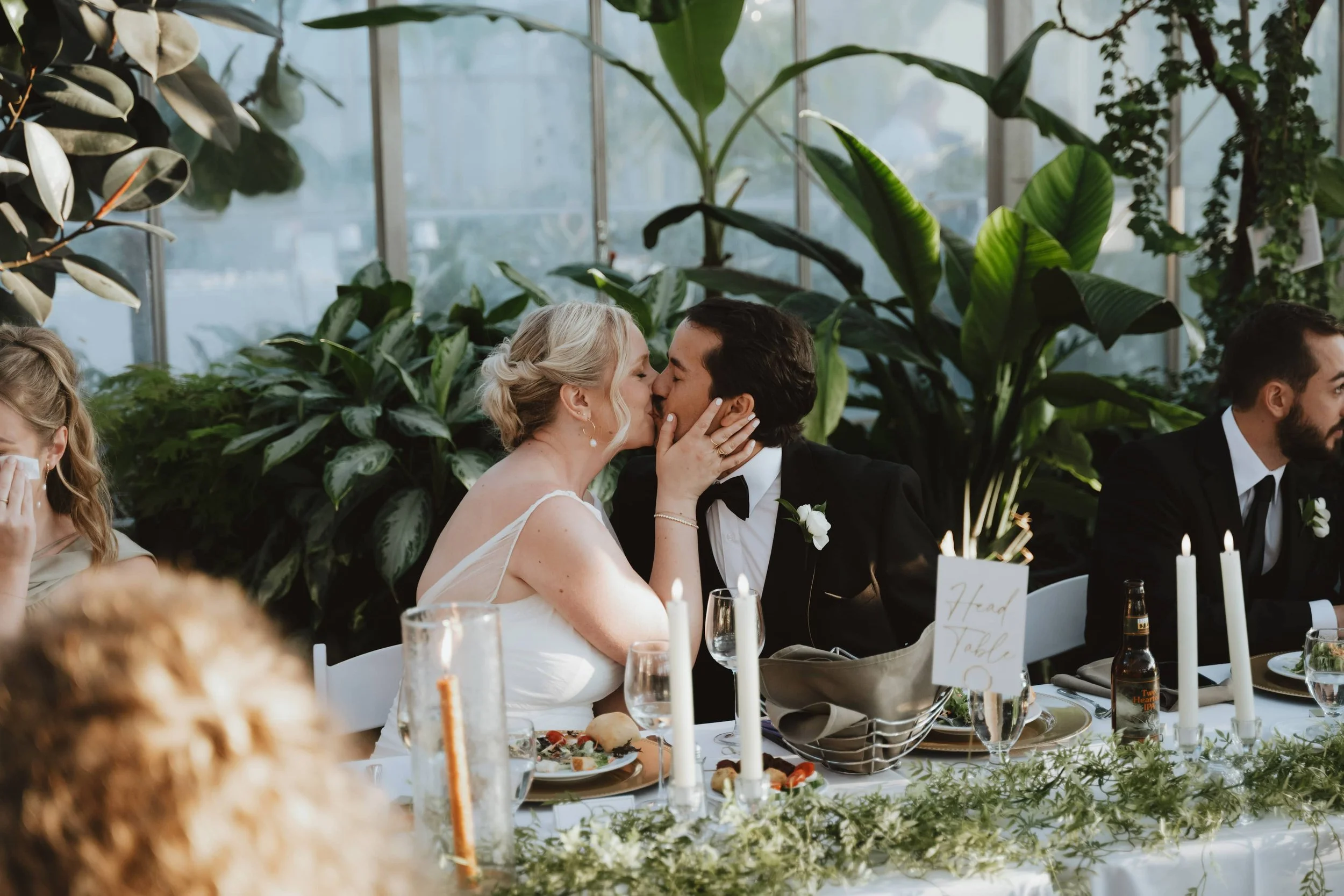 A wedding reception with a bride and groom sharing a kiss at their head table, surrounded by green plants, candles, and elegant table settings. Downtown Market Grand Rapids Michigan Wedding Venue