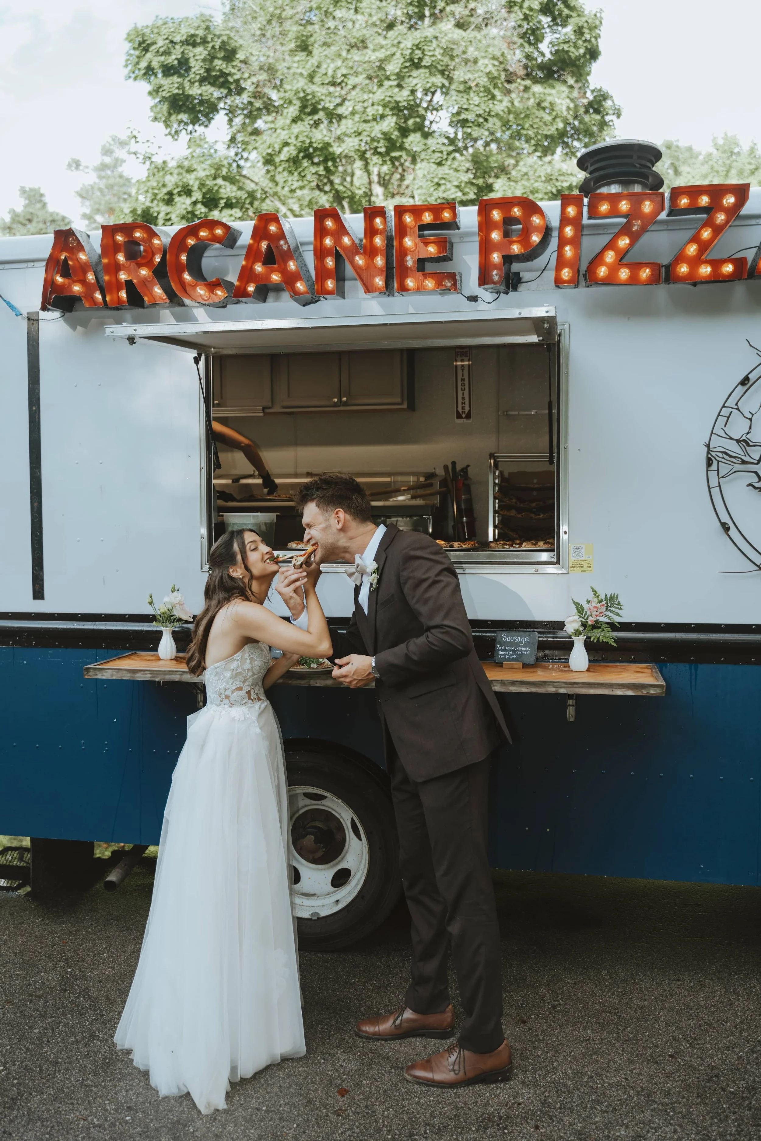 A newlywed couple sharing a slice of pizza from a food truck labeled 'Arcanepizz'