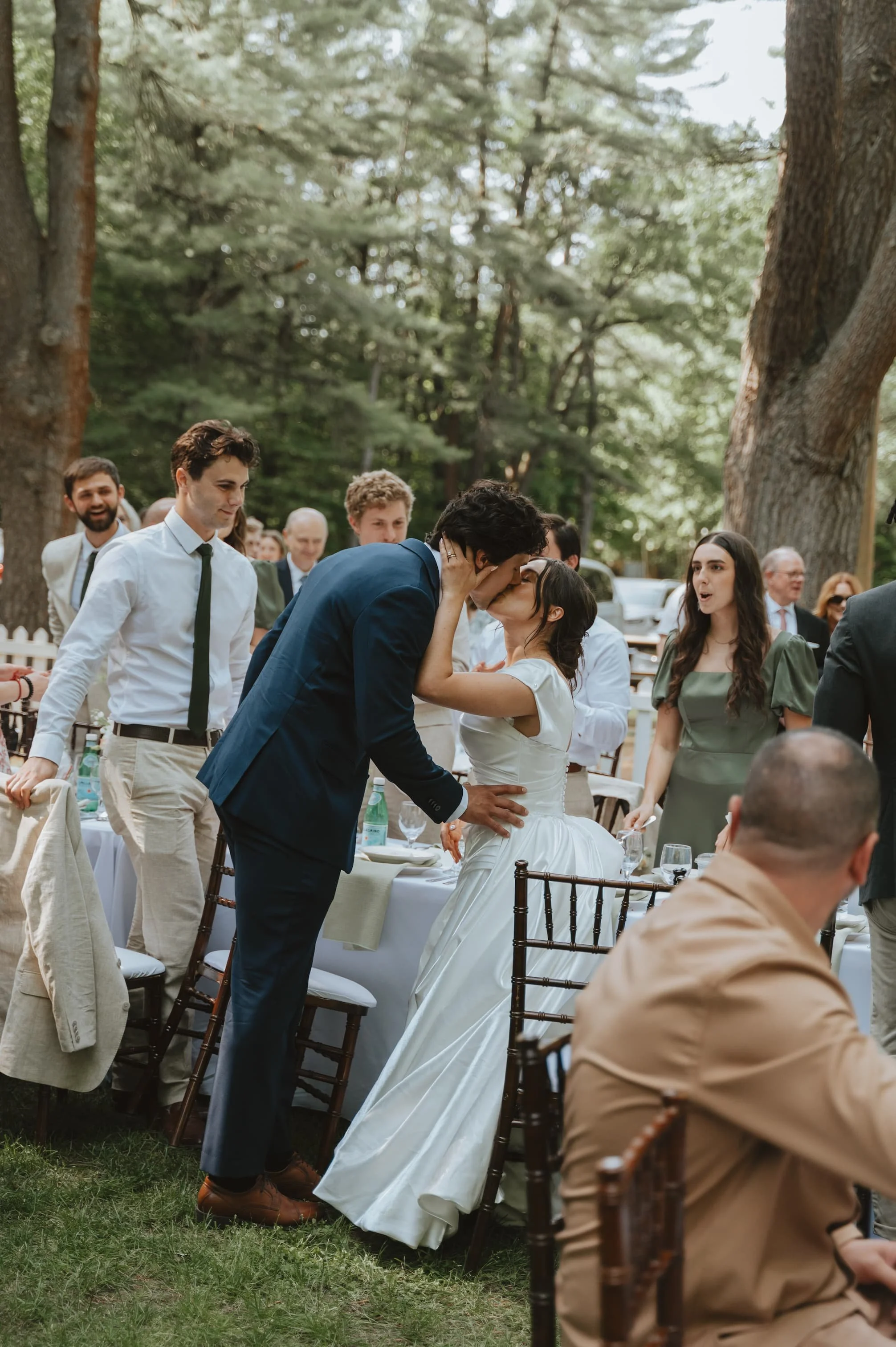 A young couple, dressed in wedding attire, shares a kiss at an outdoor wedding reception surrounded by friends and family seated at laid tables in a wooded area.