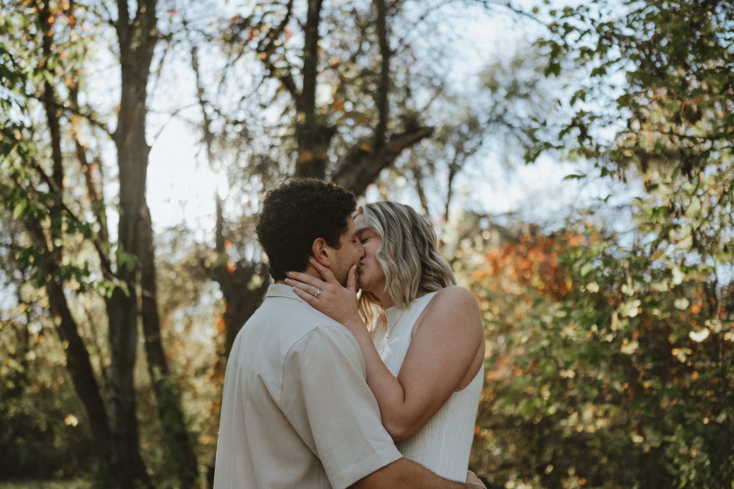 A couple sharing a kiss outdoors among trees with autumn foliage.