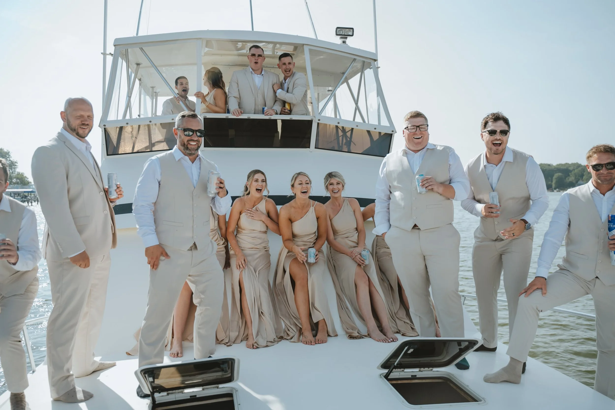 Group of men and women in beige formal attire on a boat, enjoying drinks and laughing during daytime, with some on the deck and others in the boat's upper area.