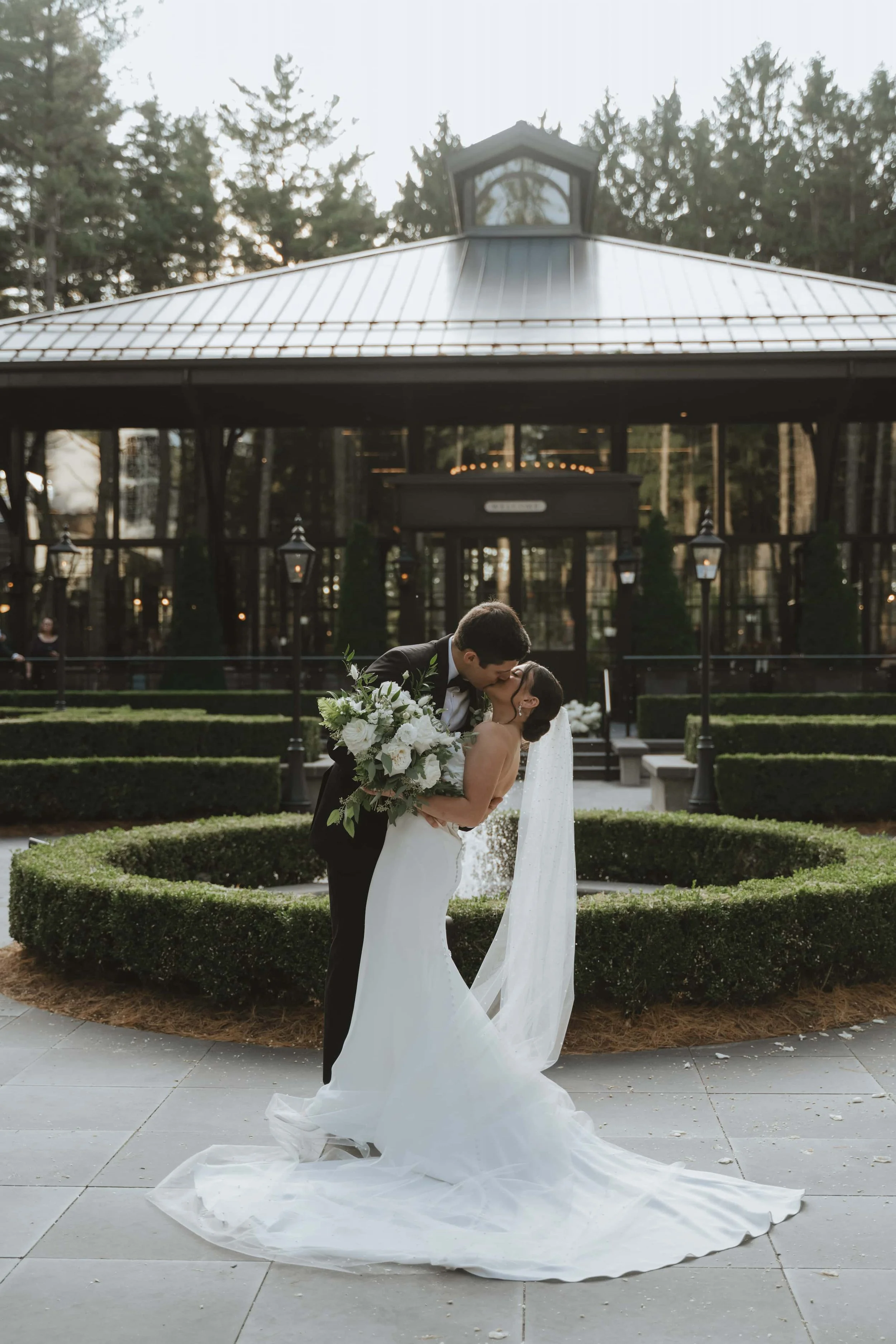 A bride and groom kissing outdoors at shepherds hollow golf course with the bride holding a large bouquet of white flowers, wedding attire, and a trailing veil.
