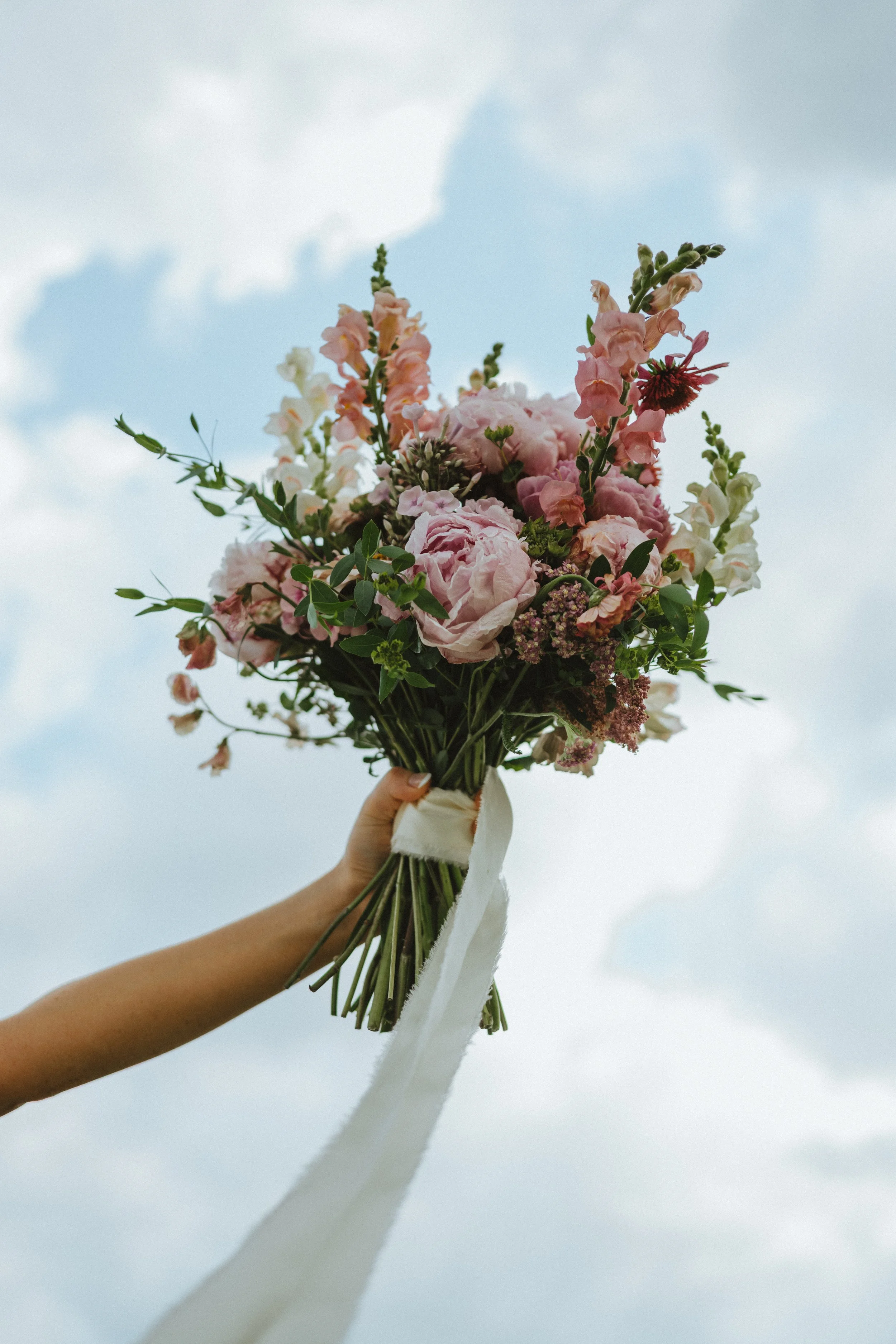 A hand holding a bouquet of pink and white flowers against a cloudy sky.