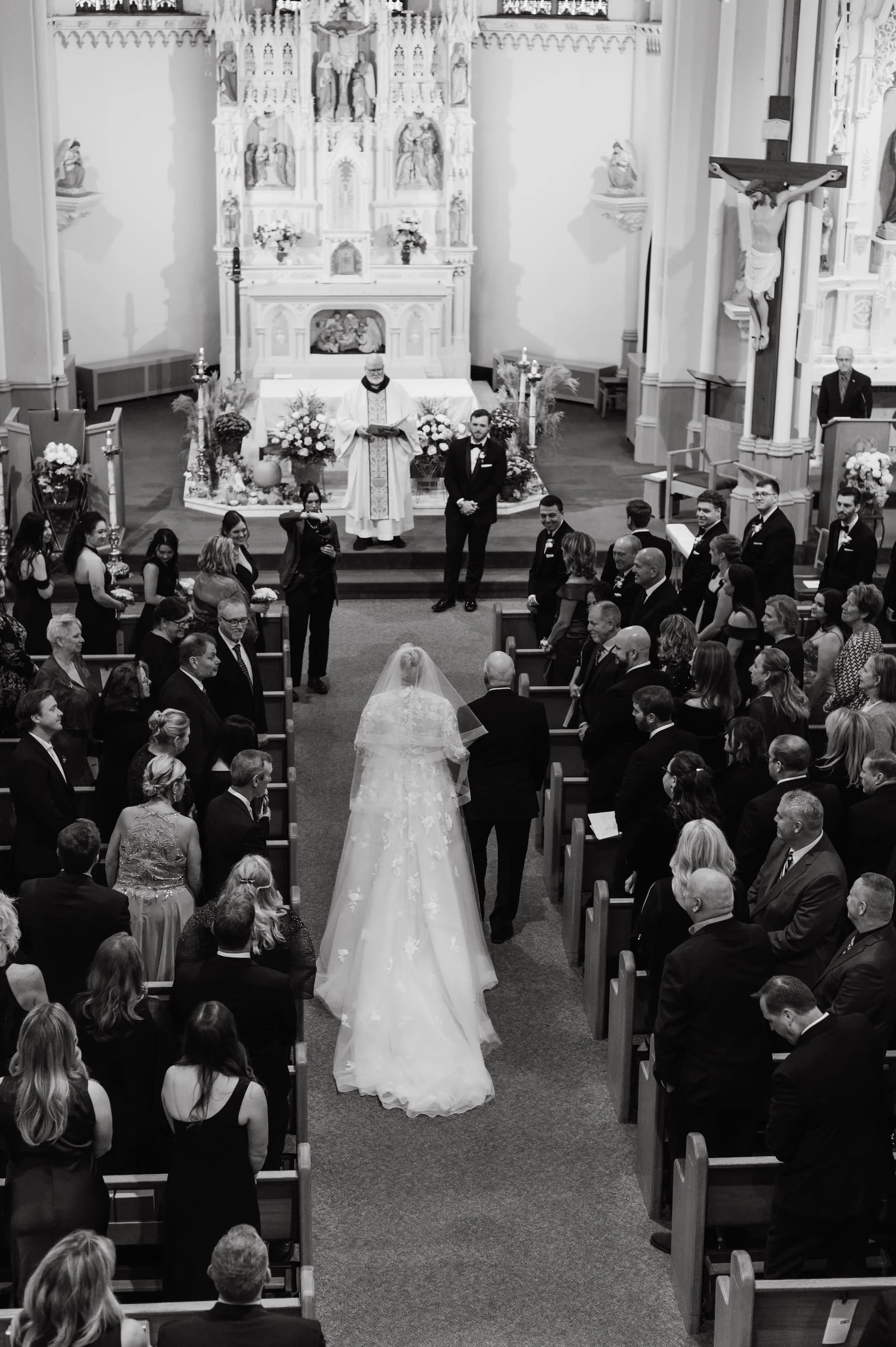 A black-and-white photo of a wedding ceremony inside a church, with a bride walking down the aisle toward the altar flanked by guests and attendants, with a priest presiding from the altar.