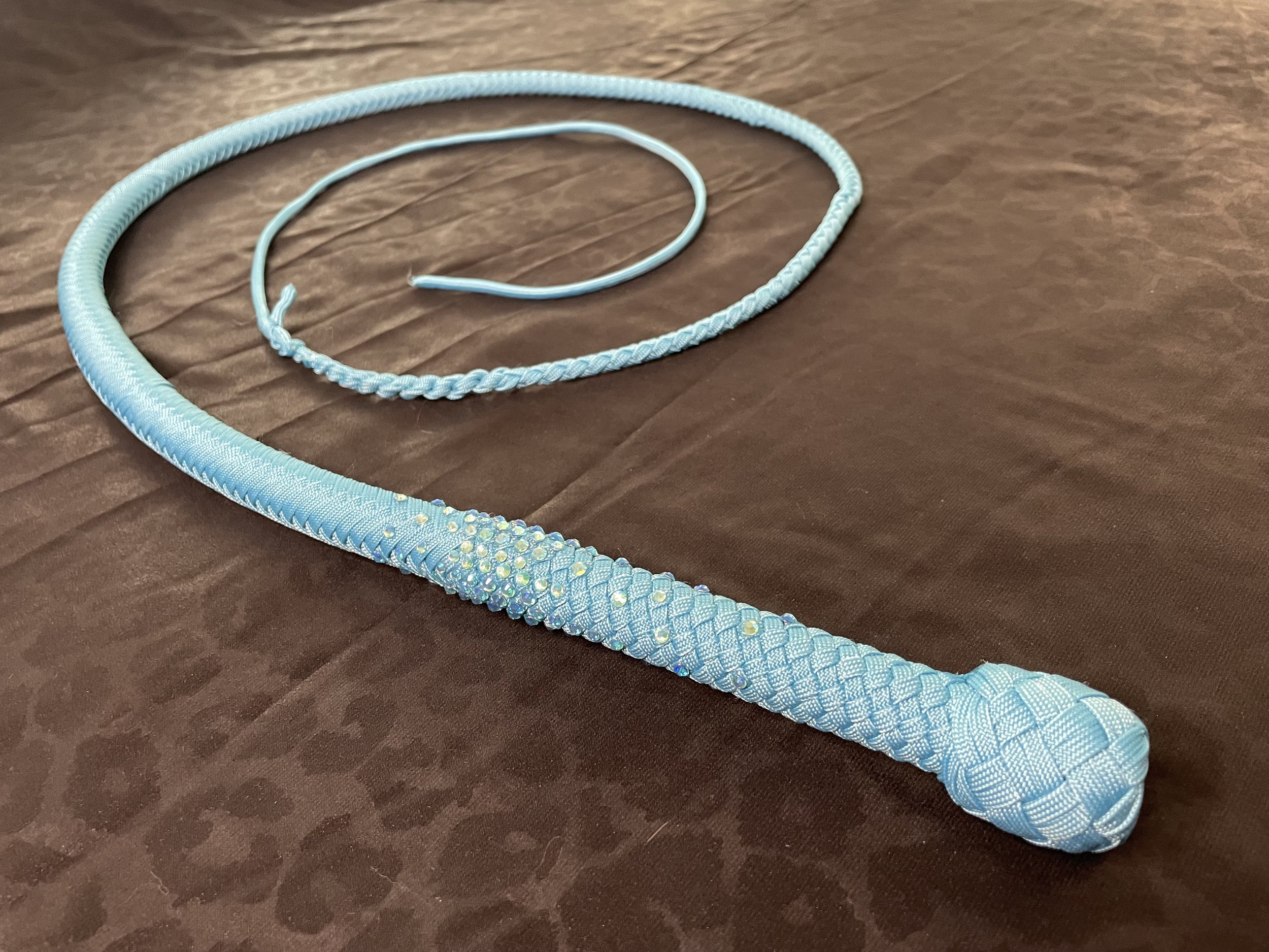 A light blue, intricately woven braided whip with a decorative handle, lying on a brown textured surface.