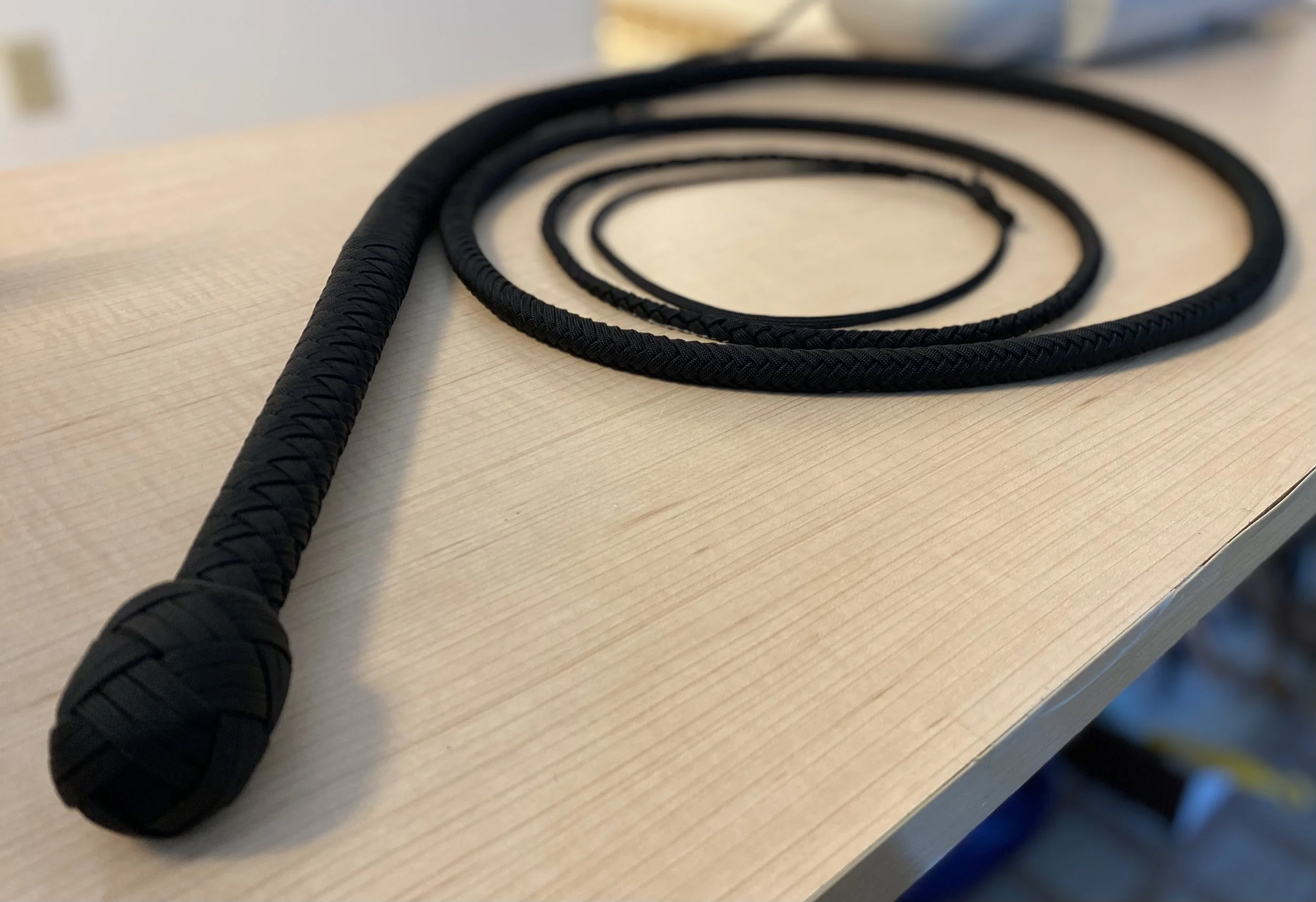 A black braided jump rope with a knotted handle lying on a light wooden surface.