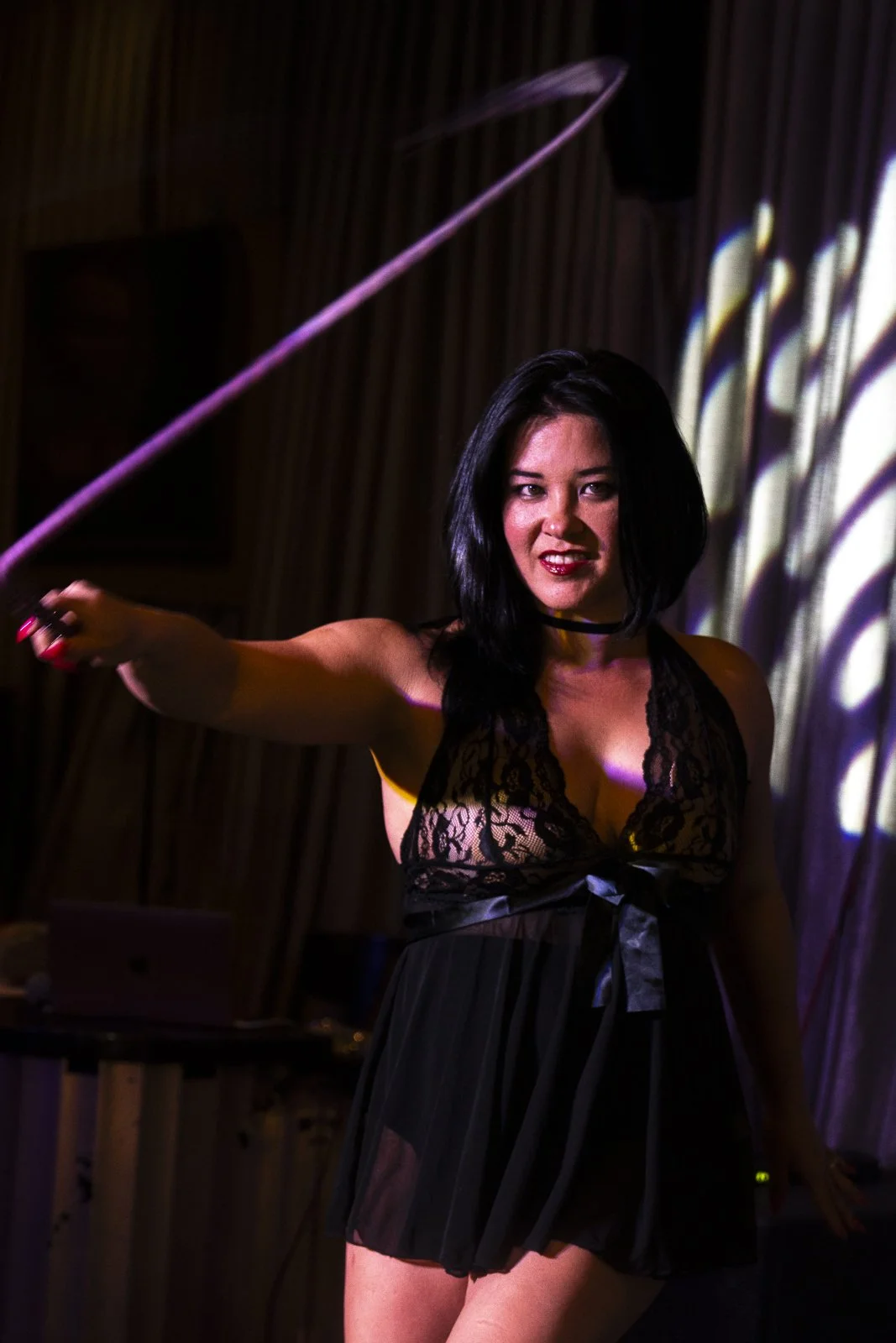A woman with black hair in a black lace revealing dress, holding a pink whip, standing against a wooden wall with colorful lighting.