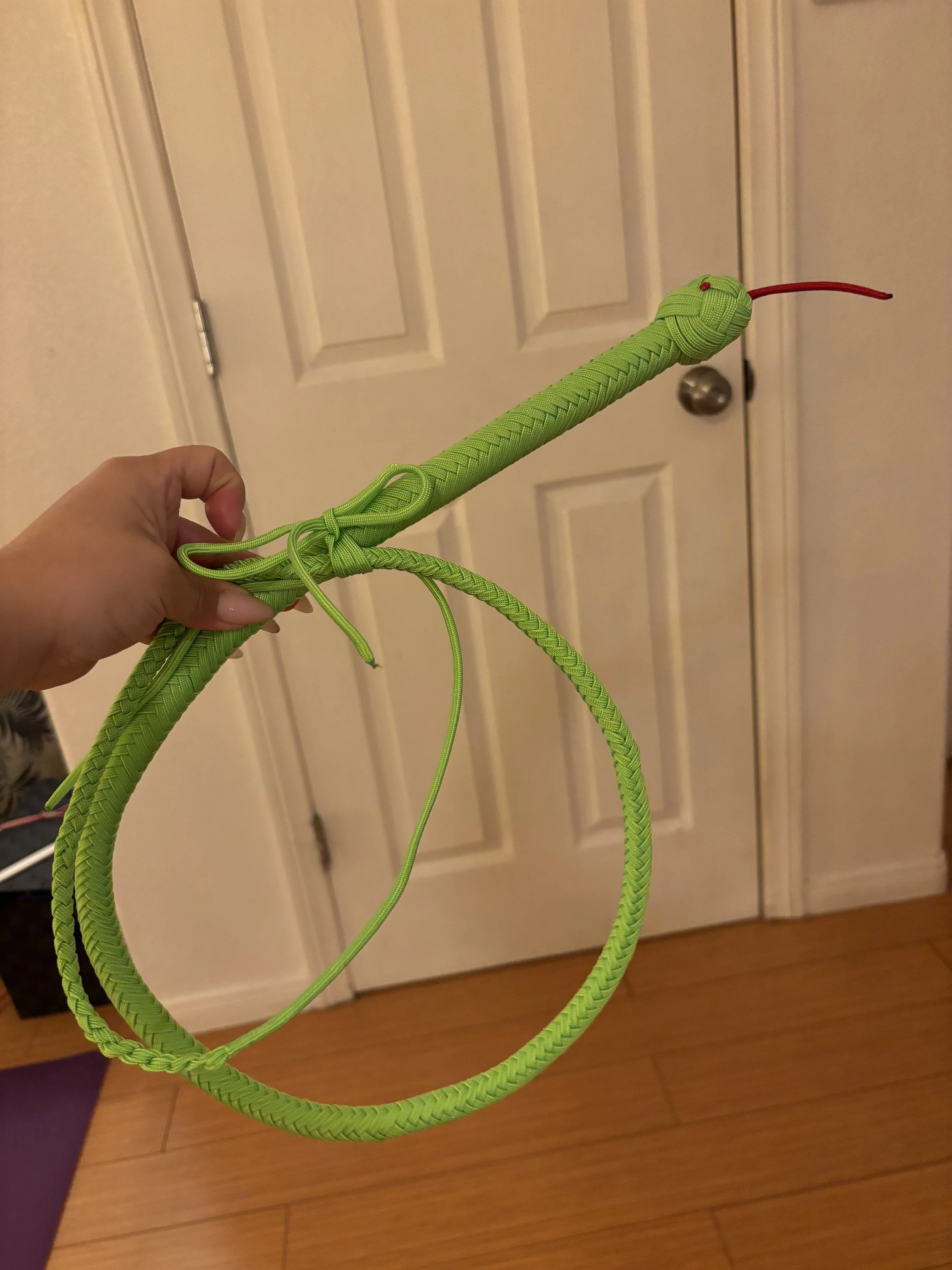 A green, snake-like craft object with a braided texture, held by a person's hand, with a red tongue-like protrusion and a small red eye, set indoors against a background of white closet doors and wooden flooring.