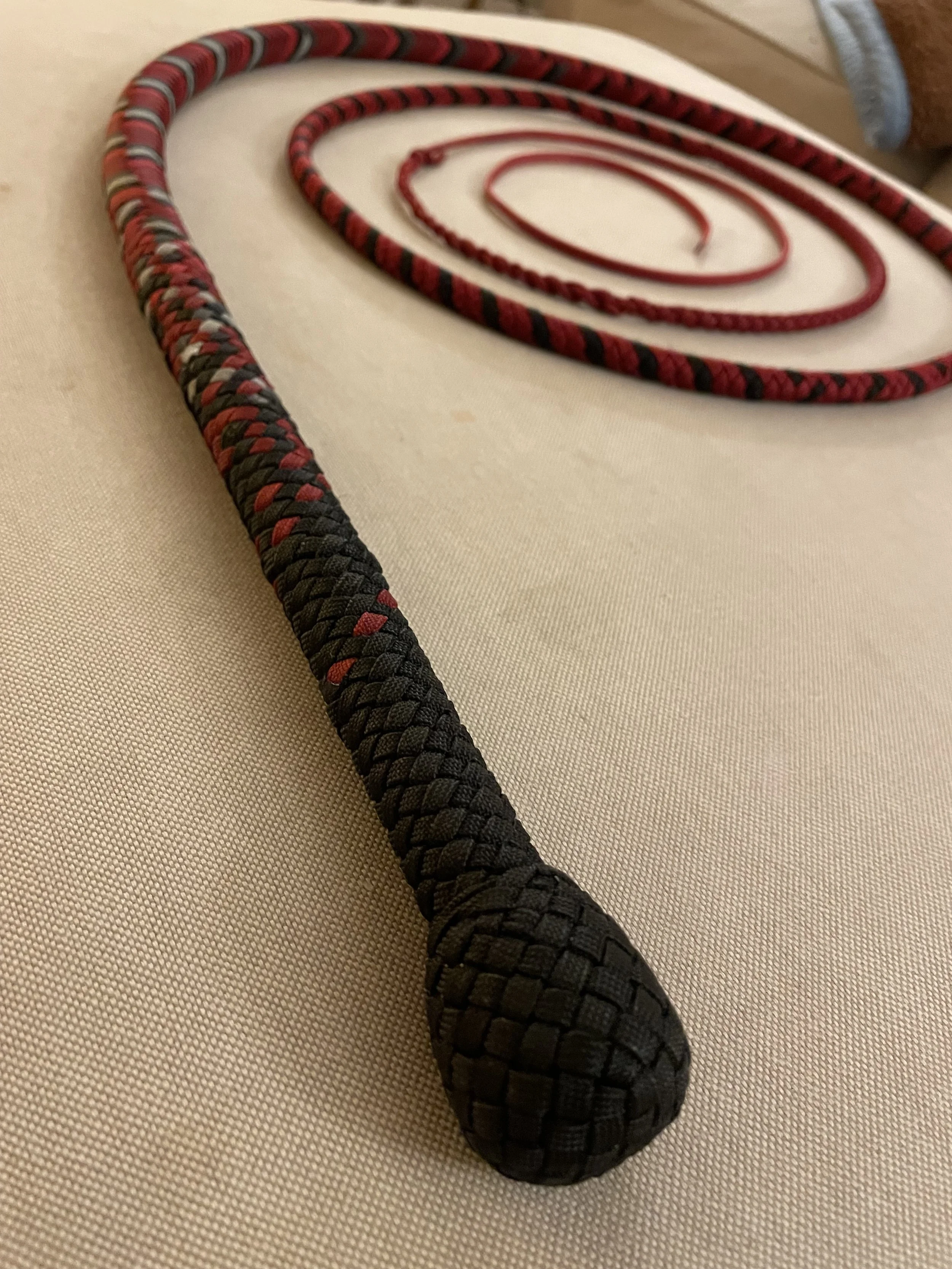 A black and red textured whip with a rounded handle lying on a beige fabric surface, with red and black jewelry shaped like a spiral in the background.