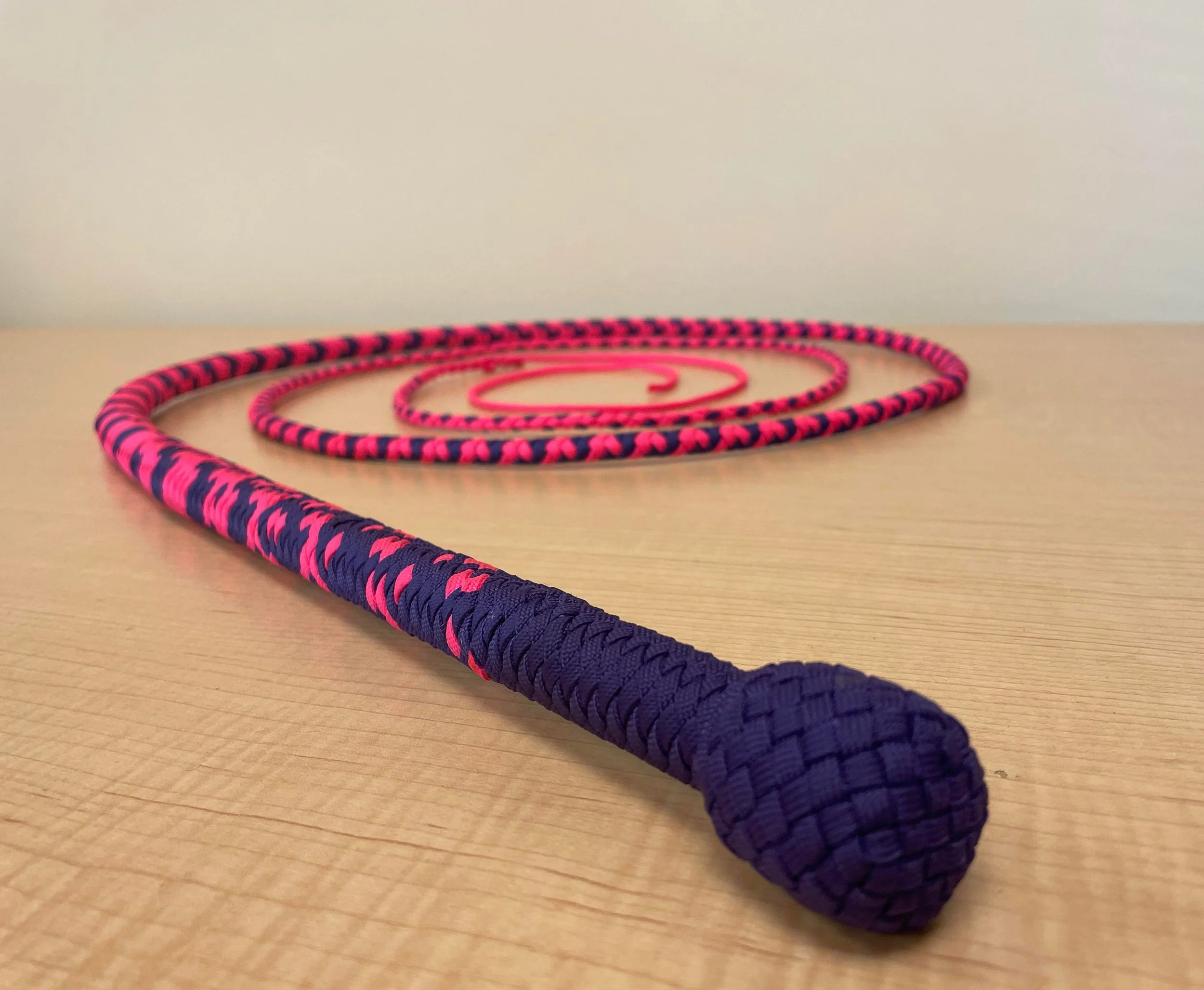 A coiled jump rope with a purple handle and a pink and navy blue woven rope, resting on a wooden surface.