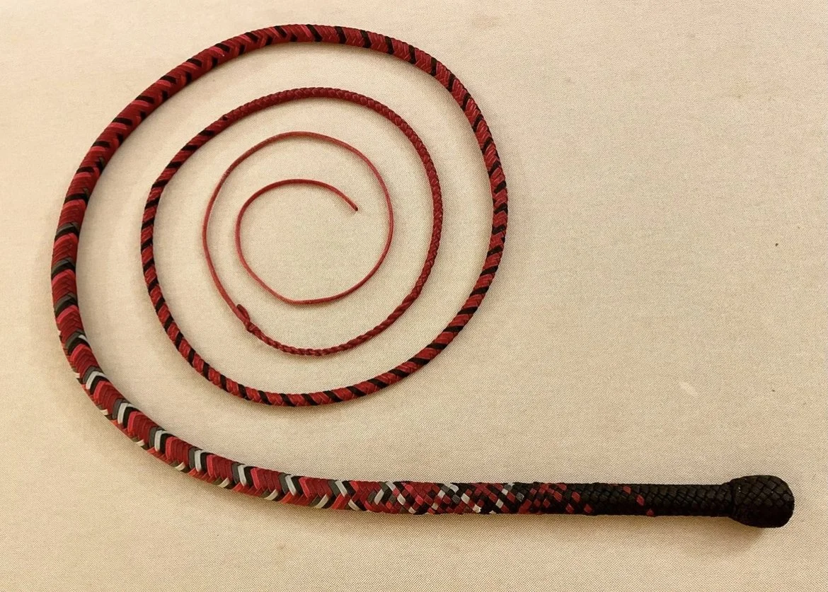 Red and black braided riding crop with a wrapped handle, placed on a beige surface.