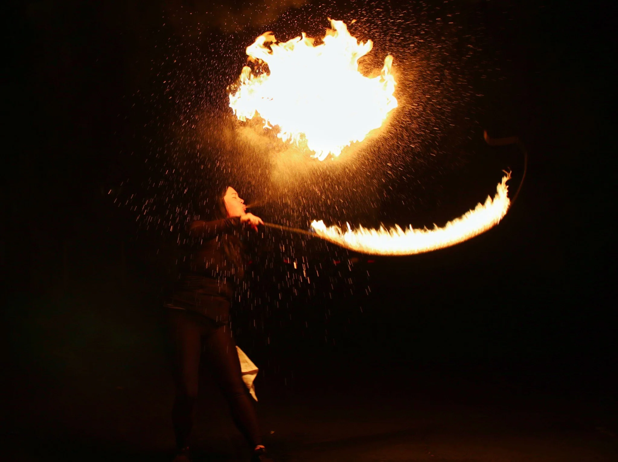 A woman performing fire dancing at night, holding a torch with flames creating a large arc of fire in the dark sky.